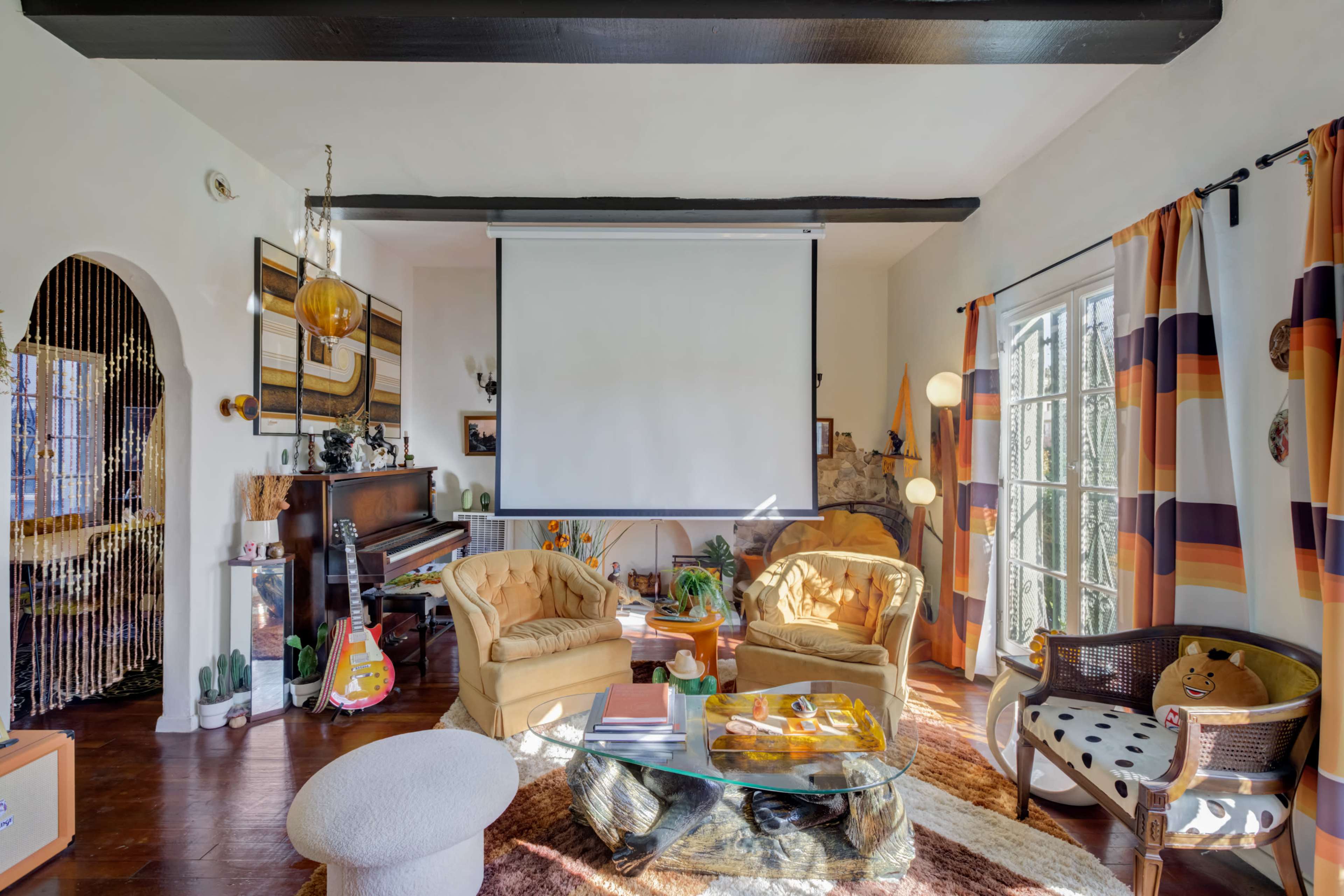A living room with two yellow armchairs, a coffee table, a large projector screen, and various decorative items, including plants and a piano.