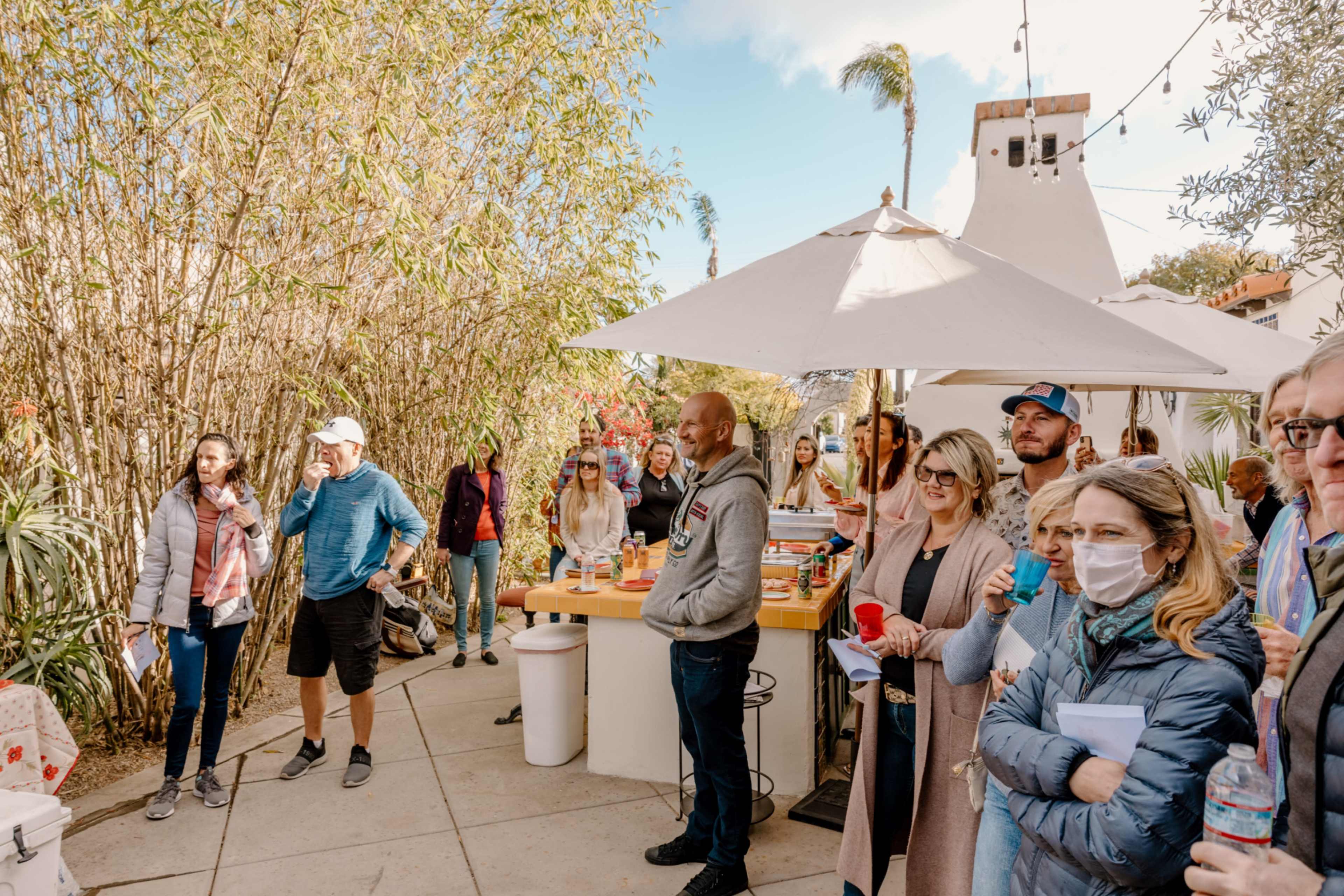 A diverse group of people gathers in a courtyard with plants and a large umbrella shading a table.