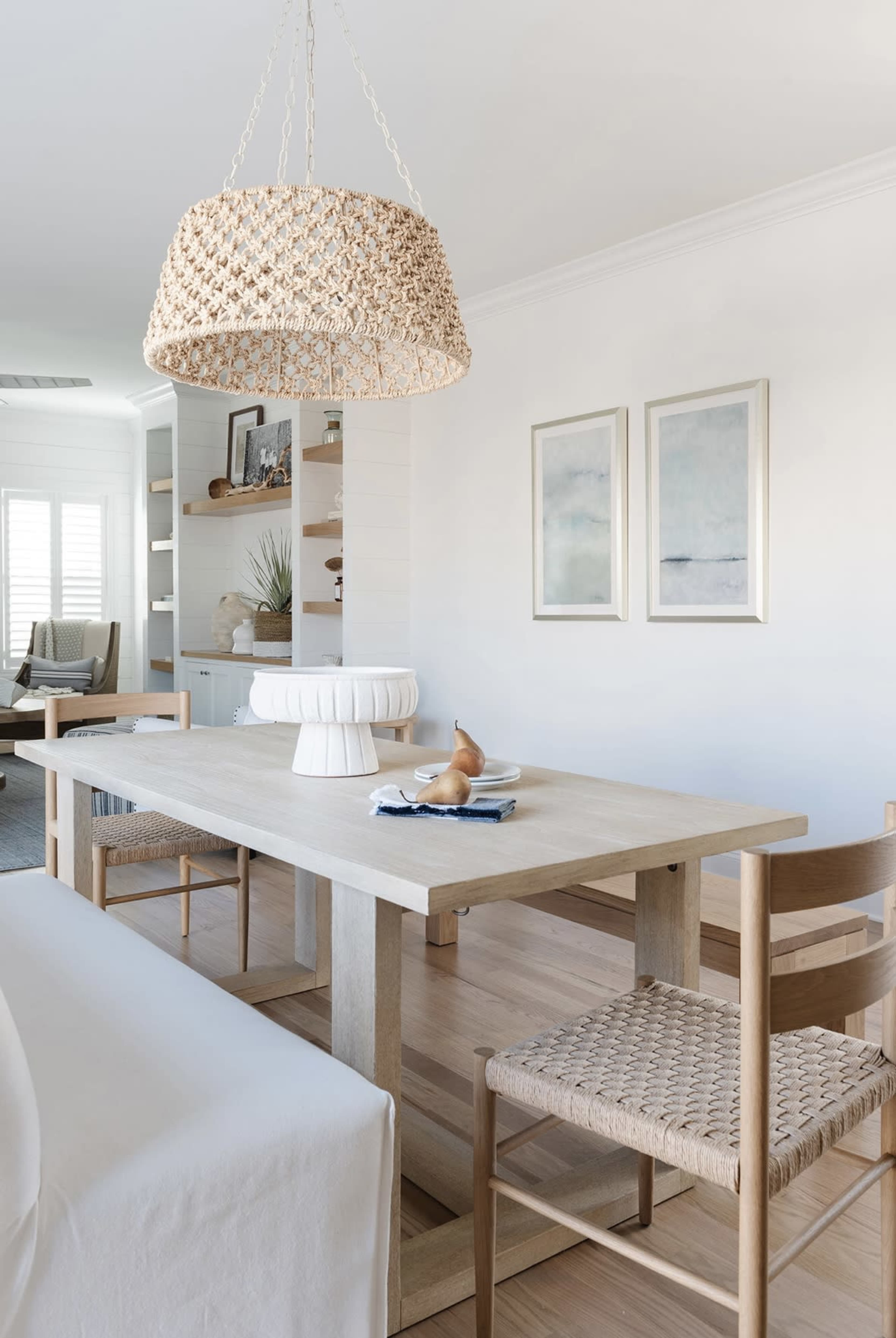 A dining area features a light wood table with a woven pendant light overhead, accompanied by two chairs and two framed artworks on the wall.
