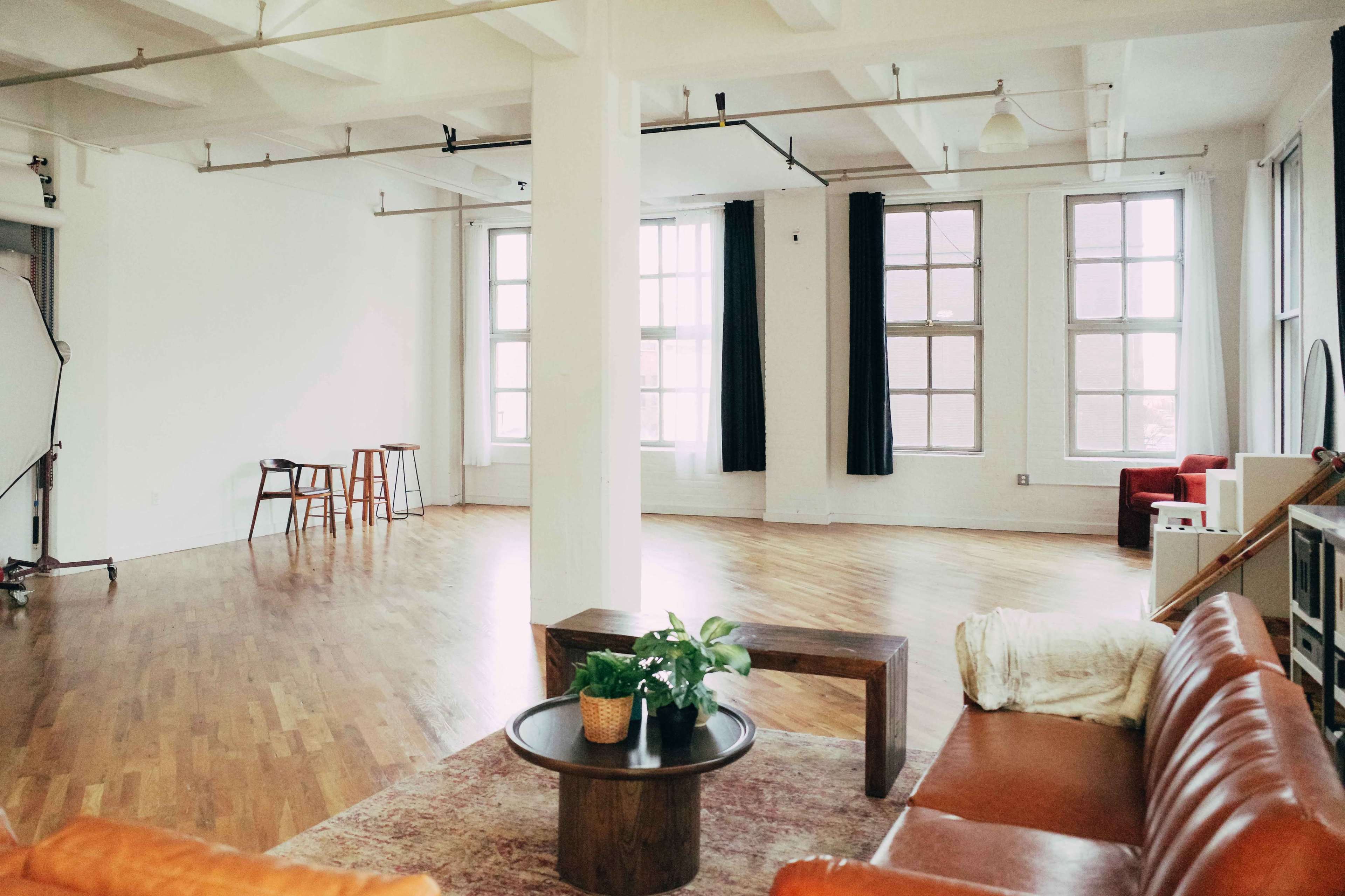 A spacious, minimalistic room with wooden flooring, large windows, and a few pieces of furniture including a brown leather sofa and a coffee table with a plant.