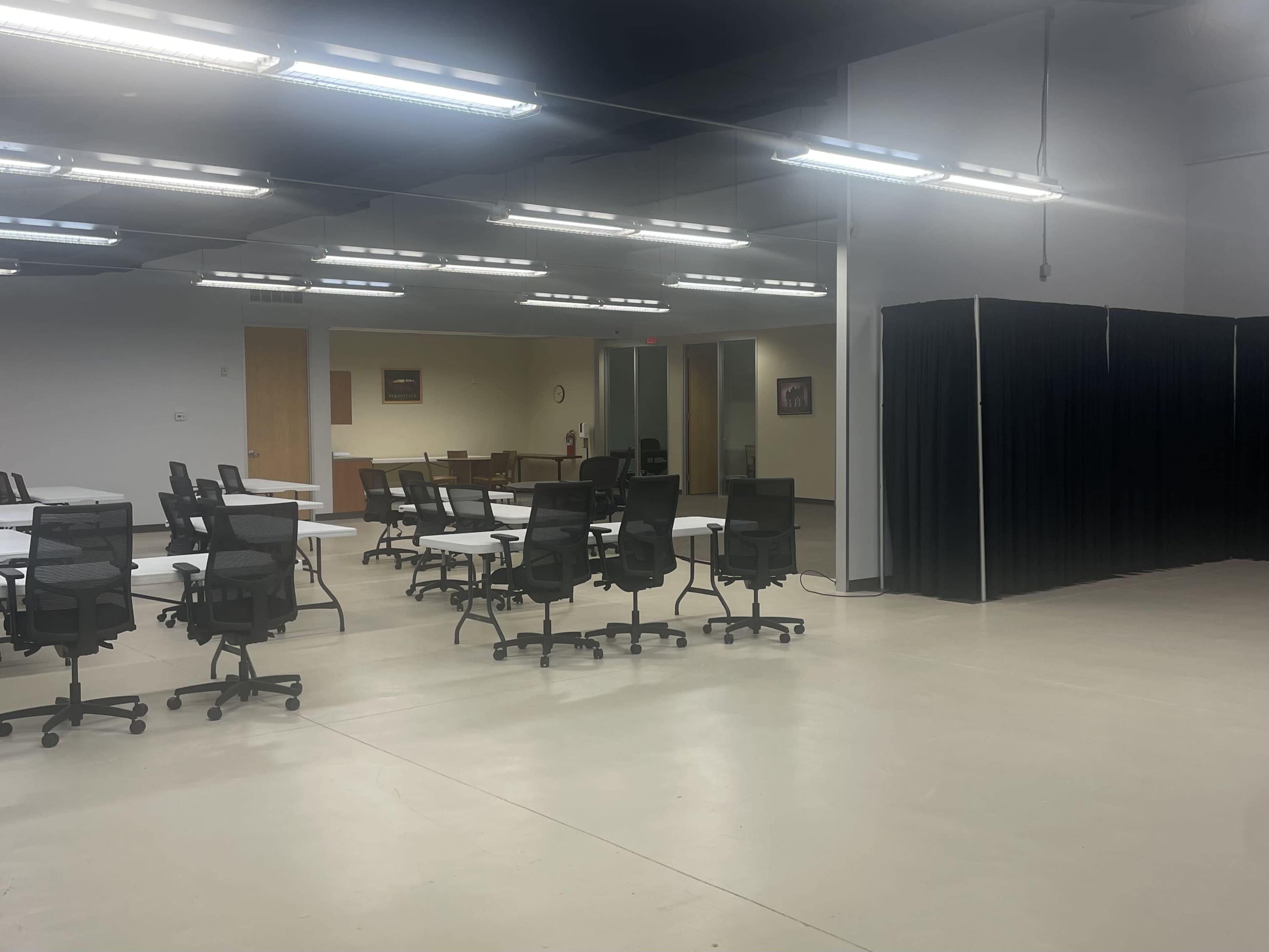 A spacious, empty office area with multiple tables and rolling chairs, alongside a partitioned section with black curtains.