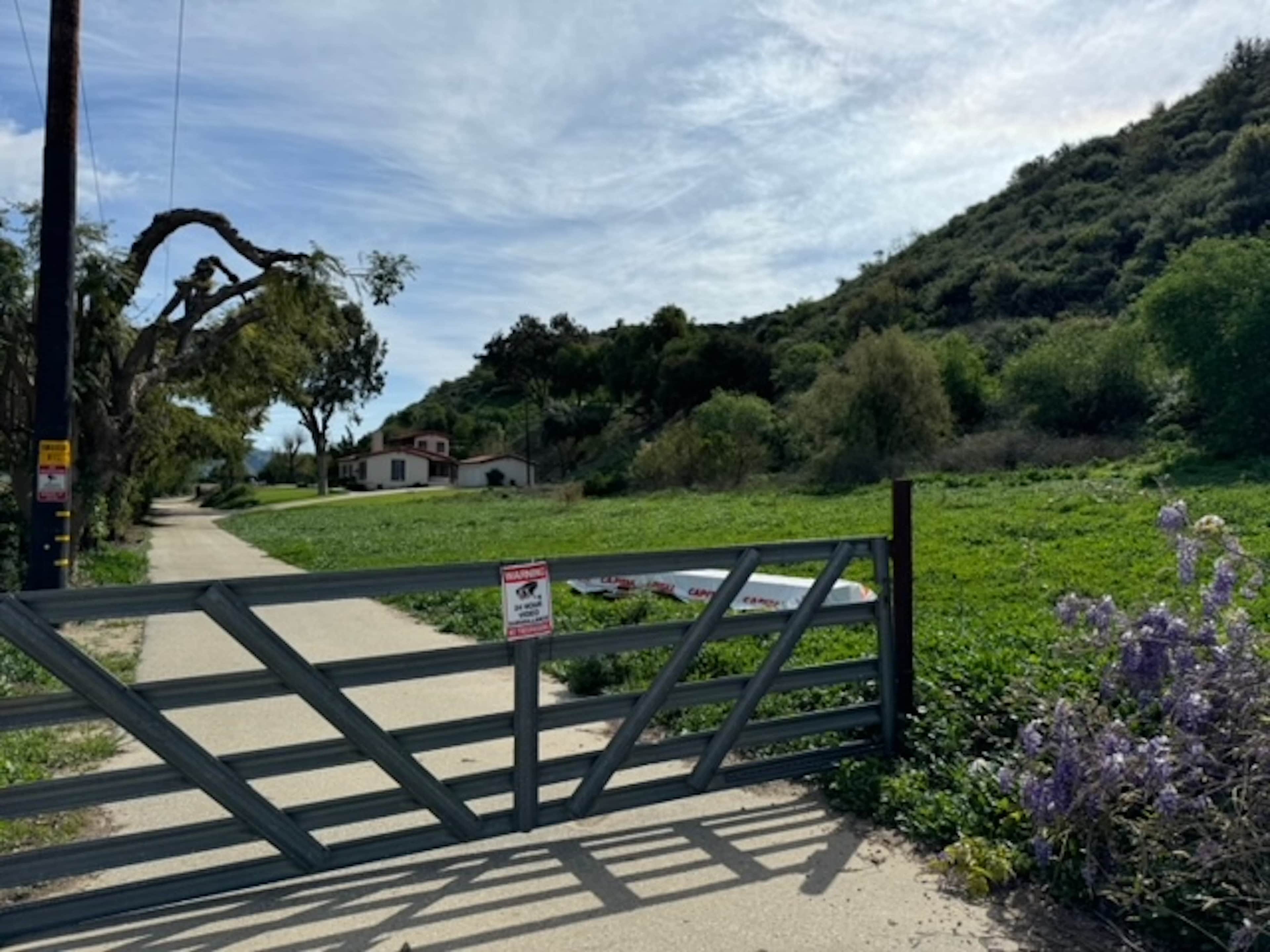 A metal gate stands at the entrance to a pathway bordered by greenery, leading to a house partially visible in the background.