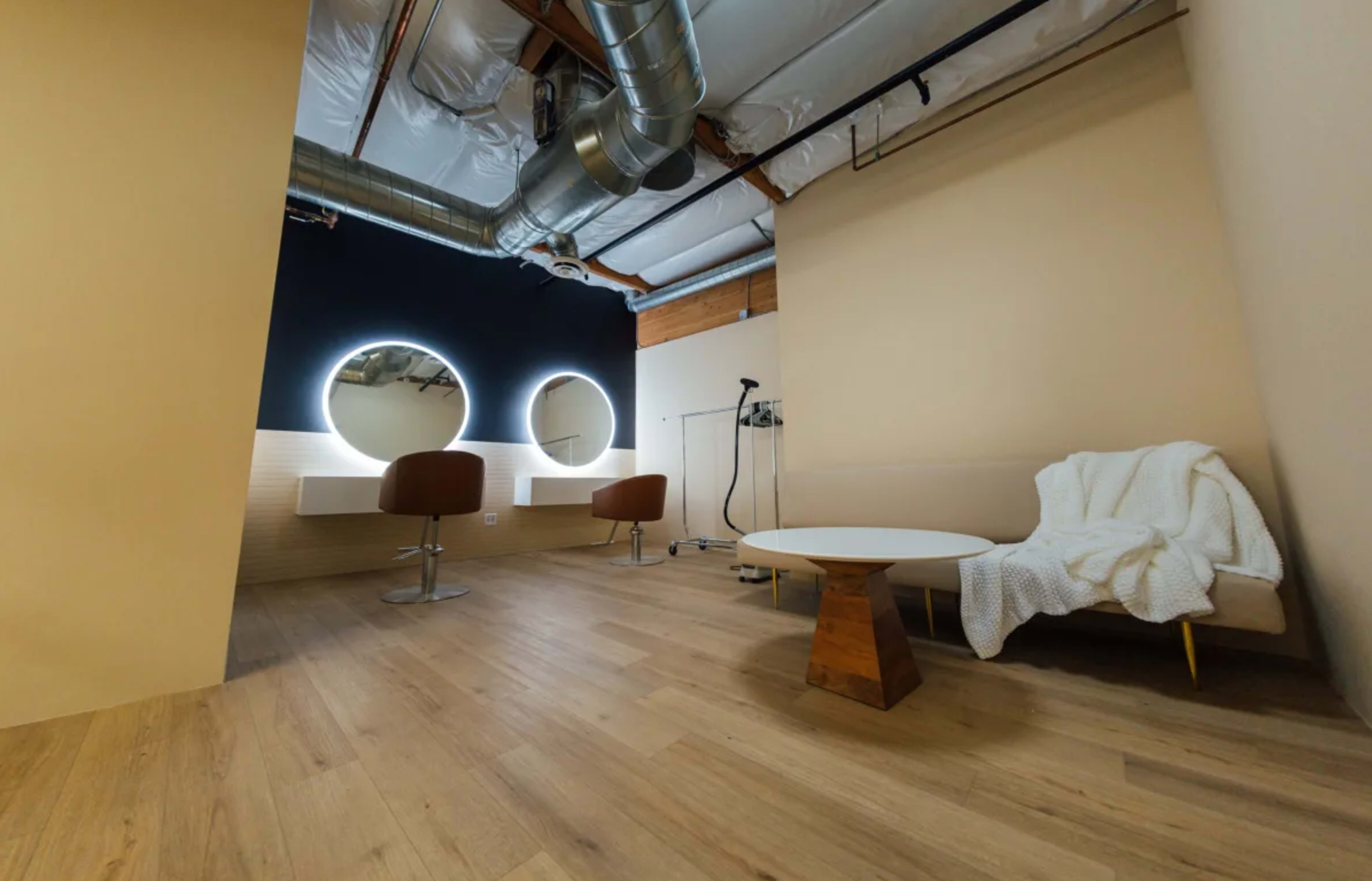 A modern salon interior with two circular mirrors, two chairs at a styling station, and a small seating area with a coffee table and a blanket.