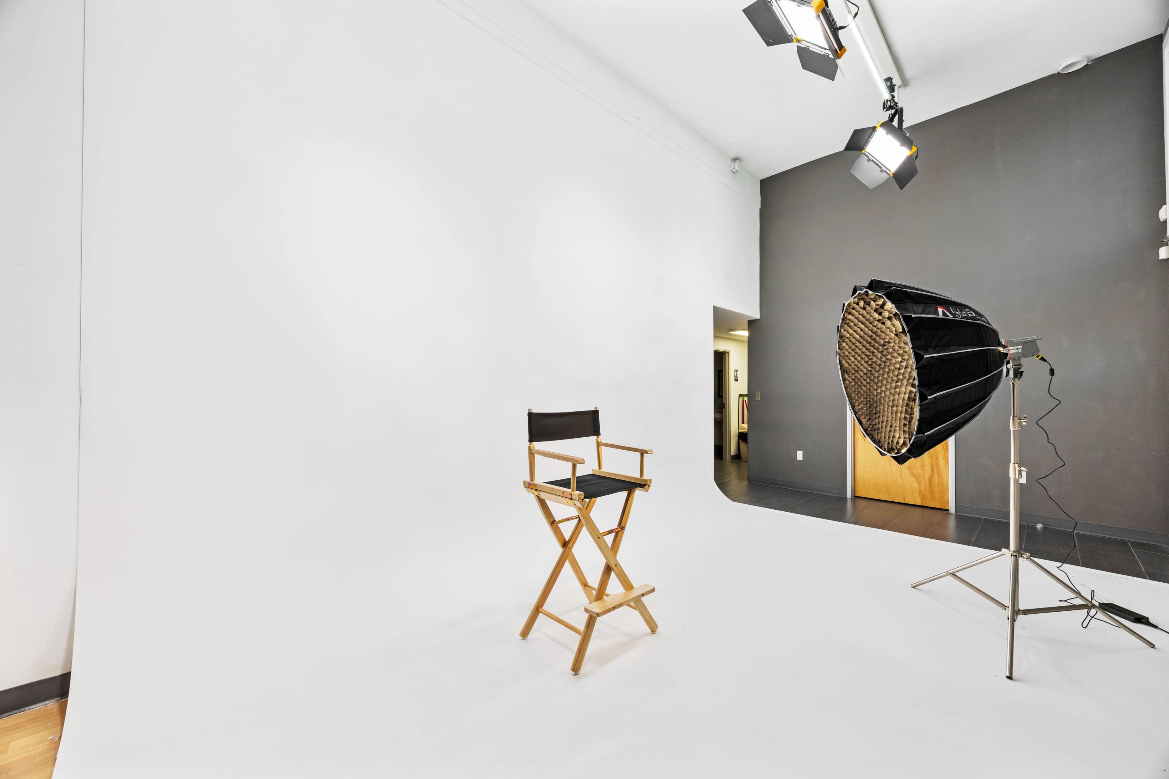 A wooden director's chair is positioned in a bright, empty photo studio with a seamless white backdrop and studio lighting equipment.