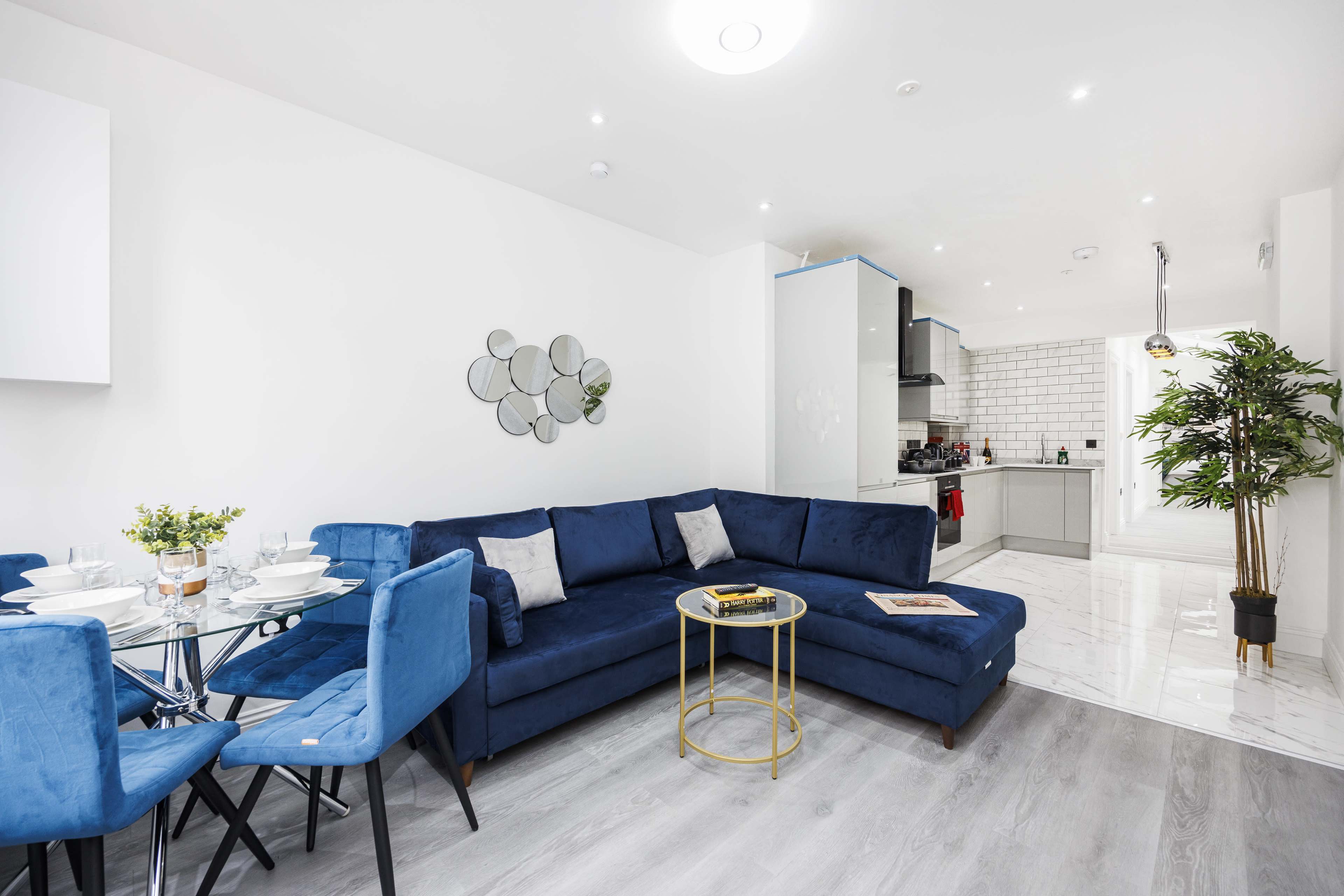 The image shows a modern living area featuring a blue sectional sofa, a round dining table set for four, and a kitchen area in the background with white cabinetry and tiled walls.