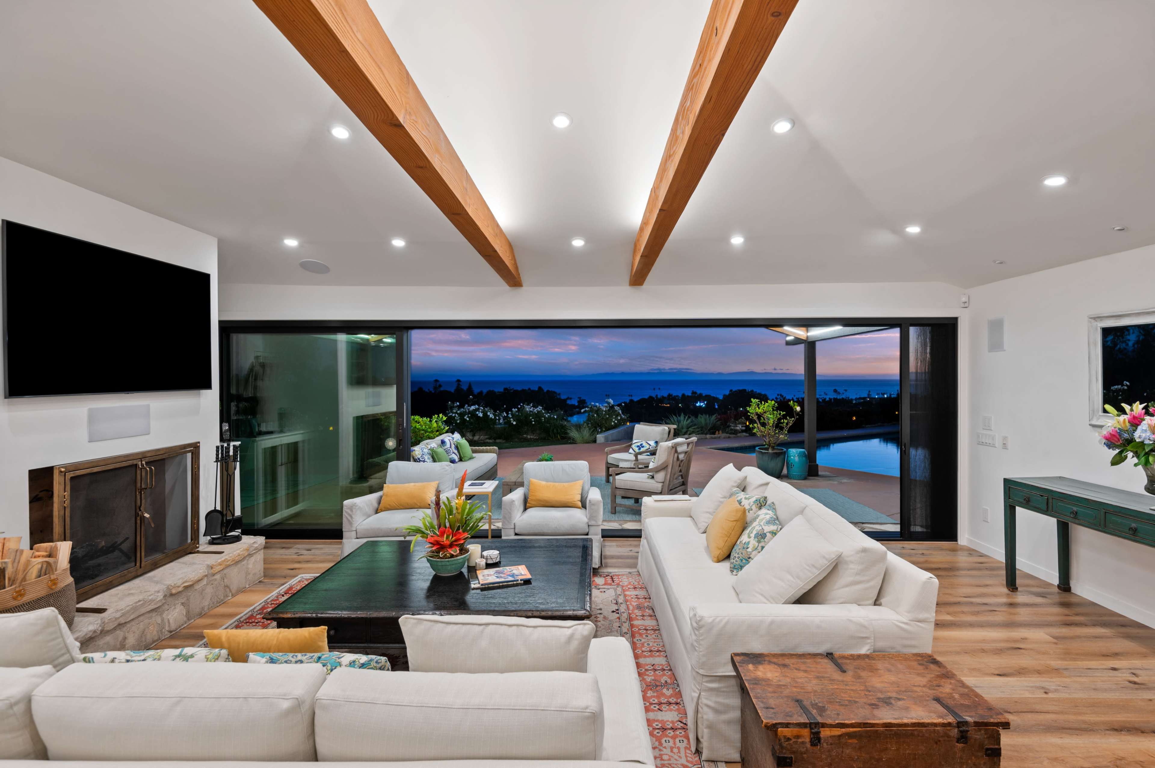 A modern living room with wooden beams, a fireplace, and large glass doors opening to an outdoor view at dusk.