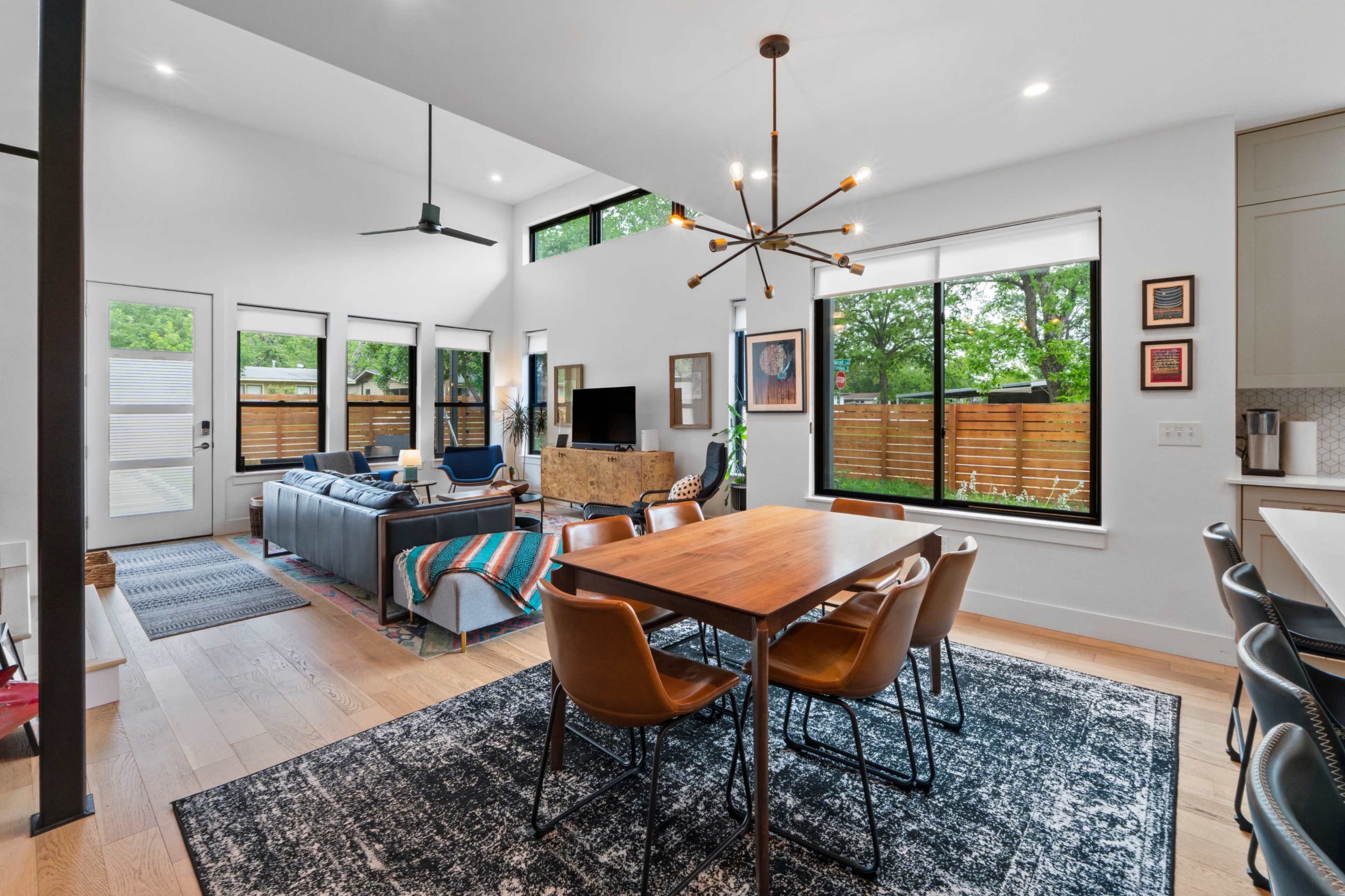 The image depicts a modern living and dining area with large windows overlooking a garden, featuring a wooden dining table, comfortable seating, and contemporary lighting fixtures.