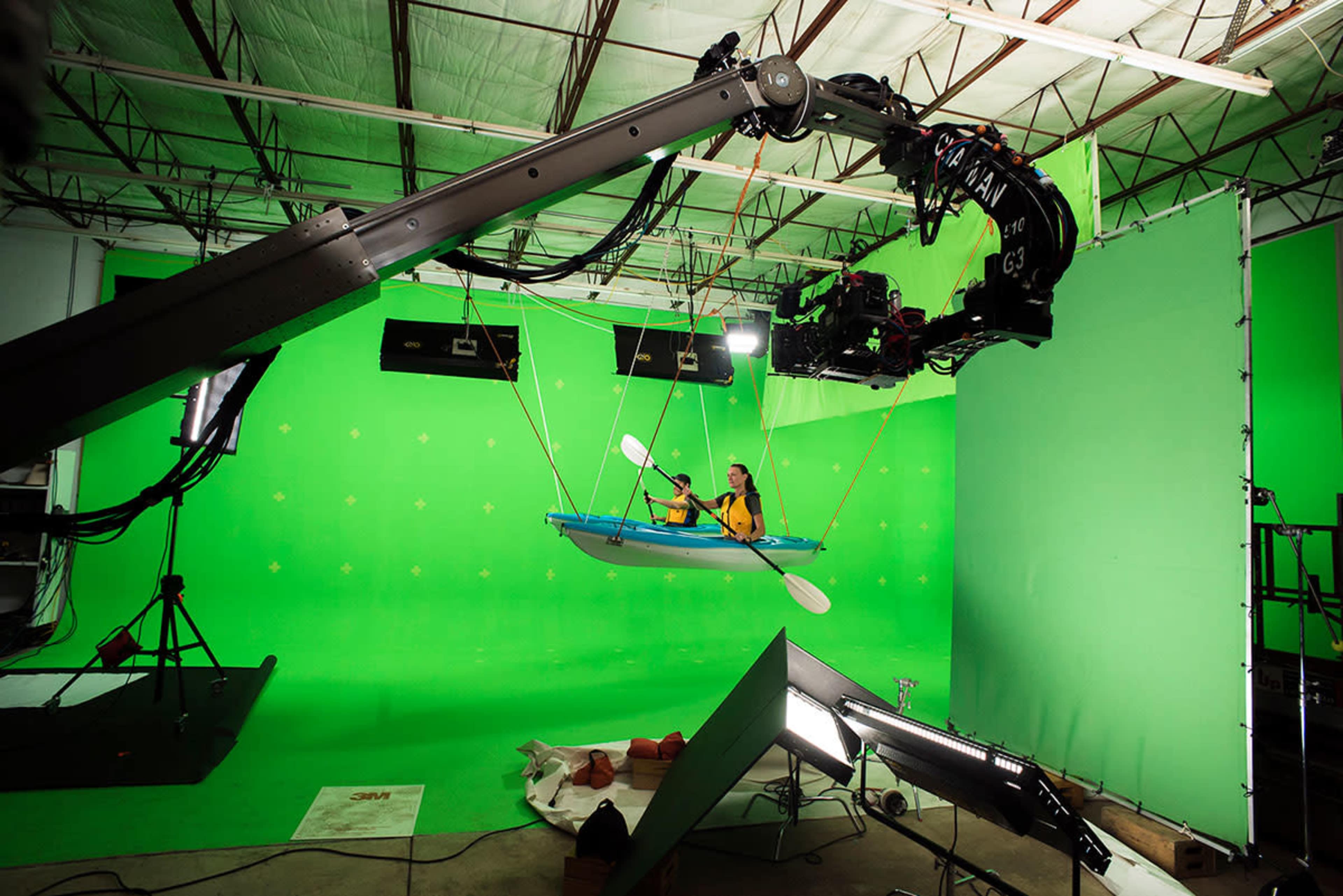 A person in a kayak is being filmed in a studio with a green screen backdrop and various lighting and camera equipment.