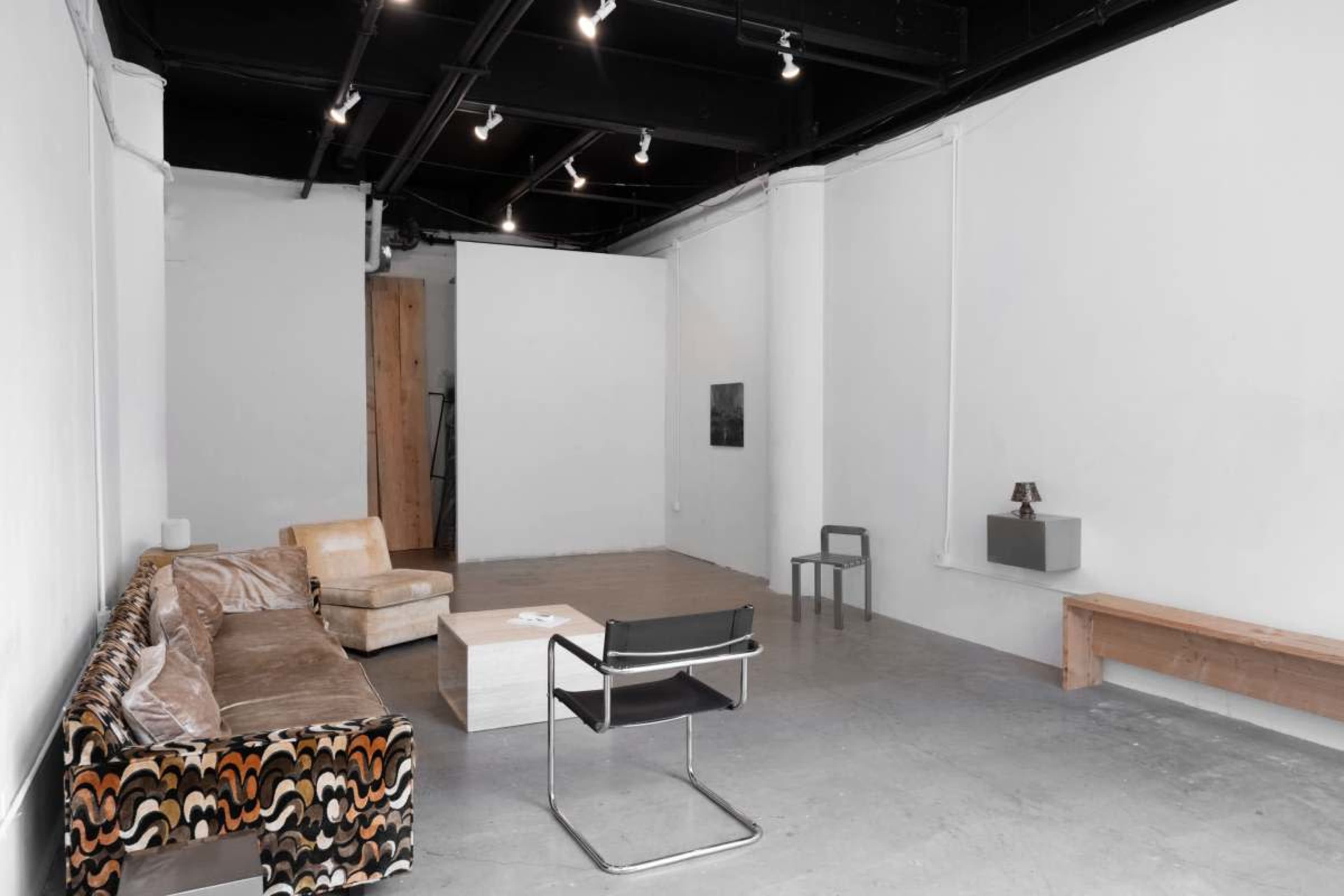 The image shows an empty gallery space with a patterned couch, a simple coffee table, a black chair, and a wooden bench against the walls.
