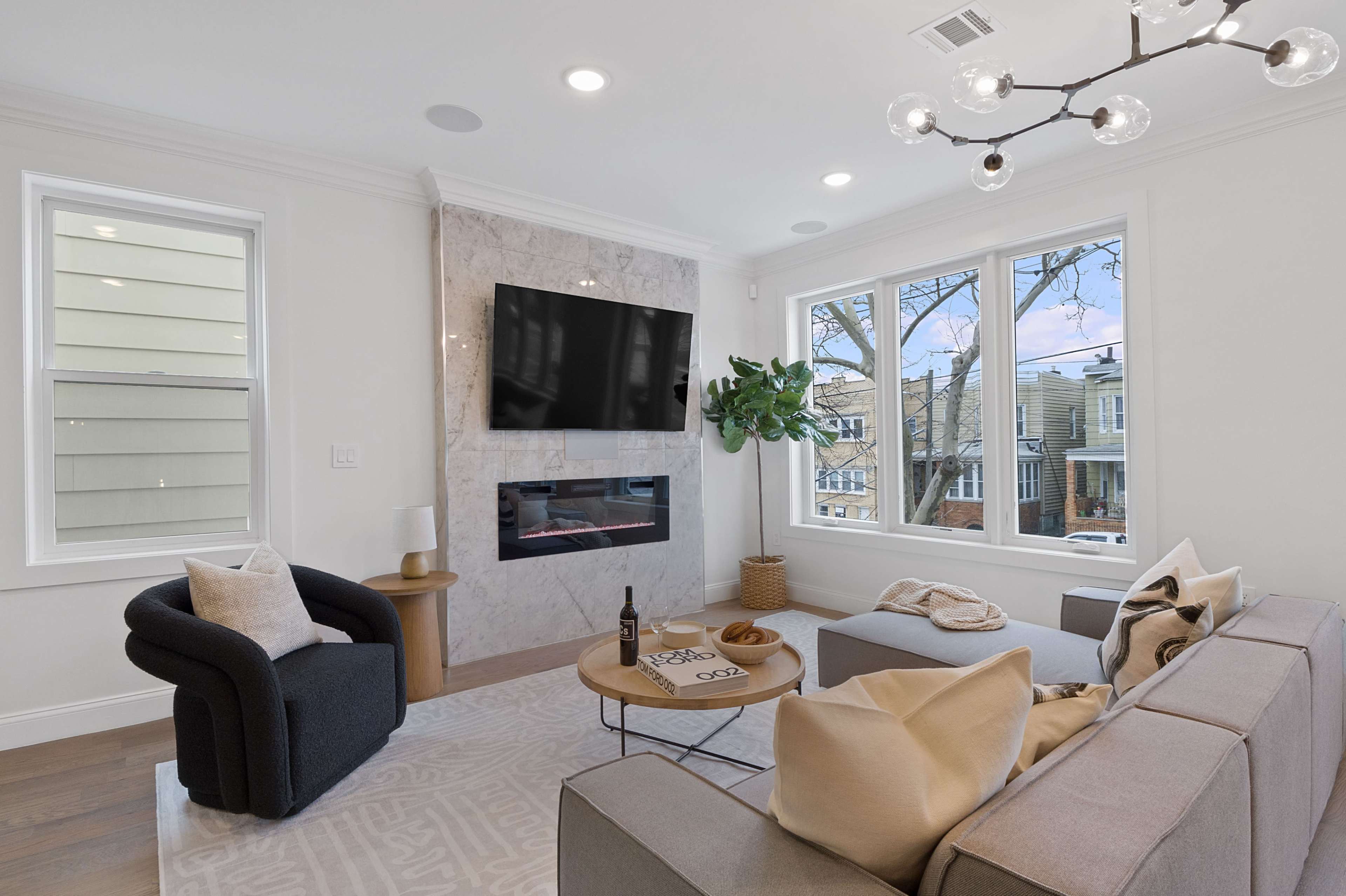 A modern living room features a large wall-mounted television, a minimalist fireplace, a dark chair, a sectional sofa with cushions, and a round coffee table.