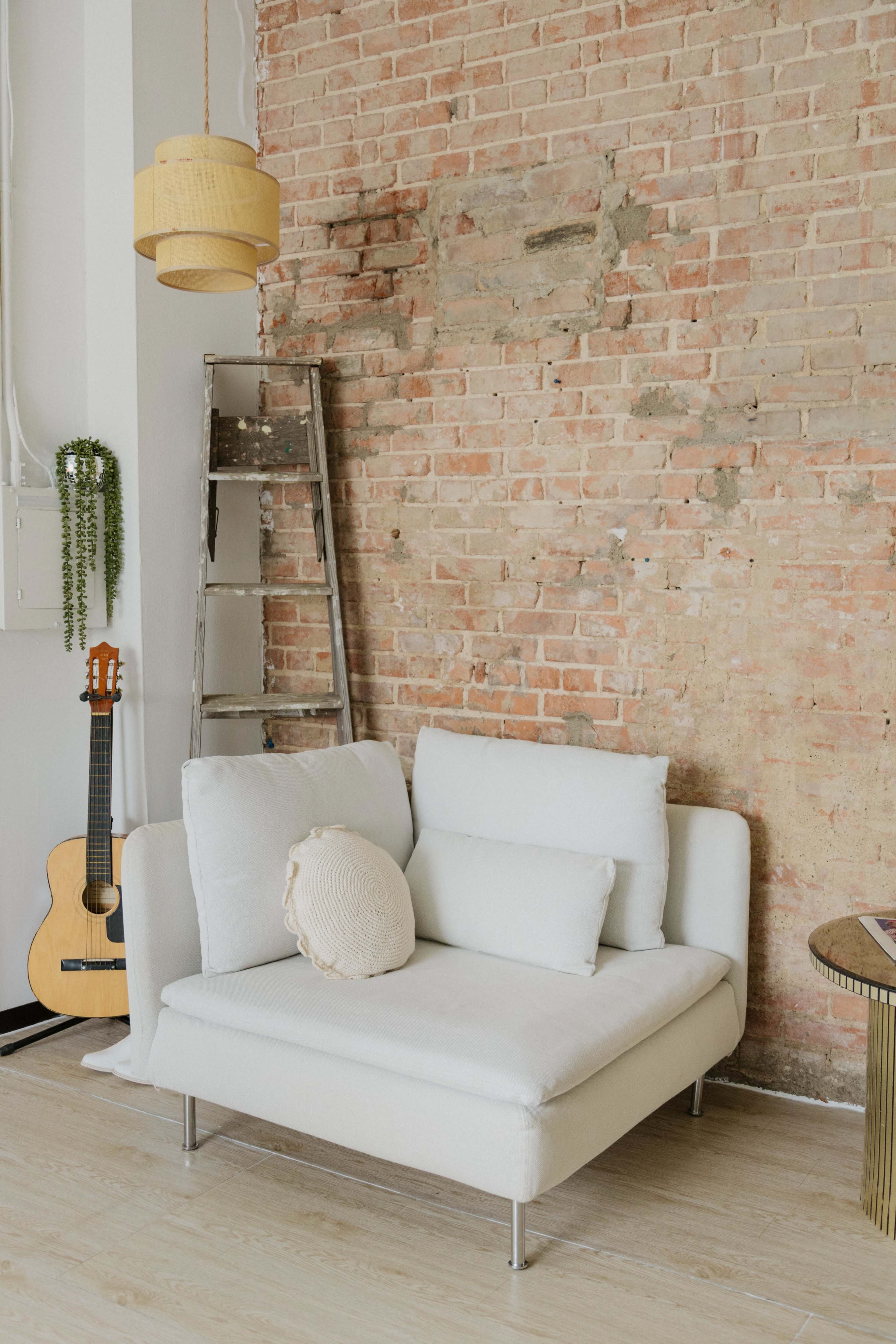 A white sofa with decorative cushions is positioned against a rustic brick wall, accompanied by a ladder, an acoustic guitar, and a pendant lamp.