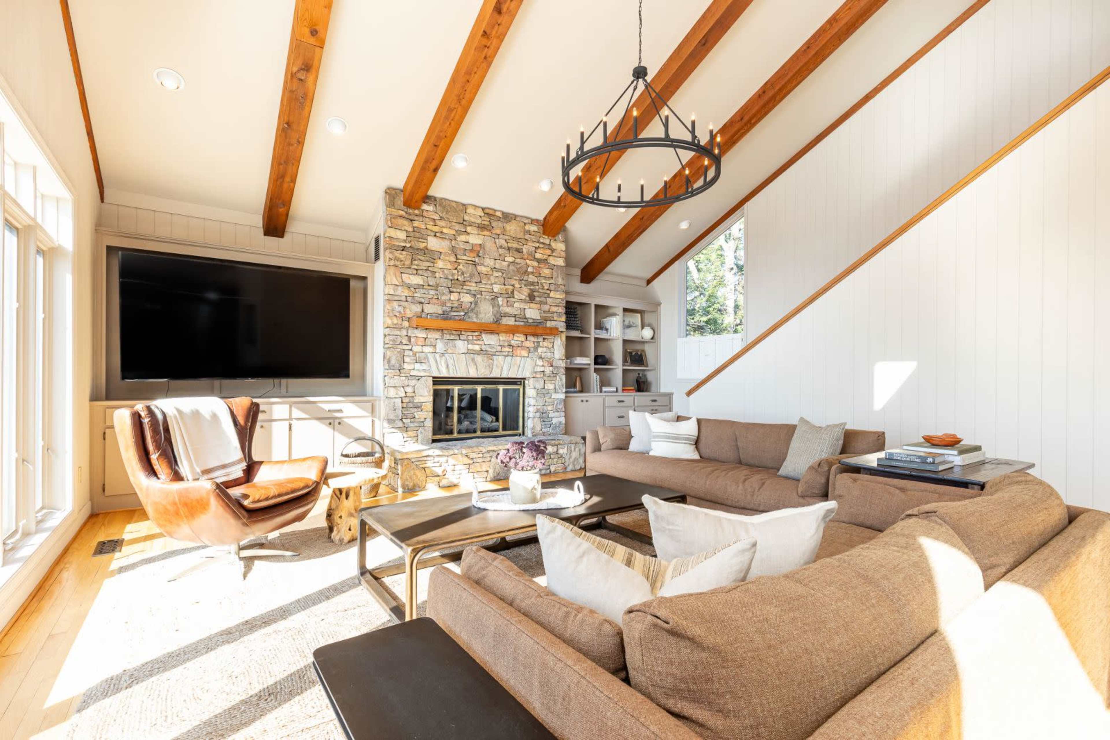 A spacious living room features a stone fireplace, a large sectional sofa, and wooden beams on the ceiling.
