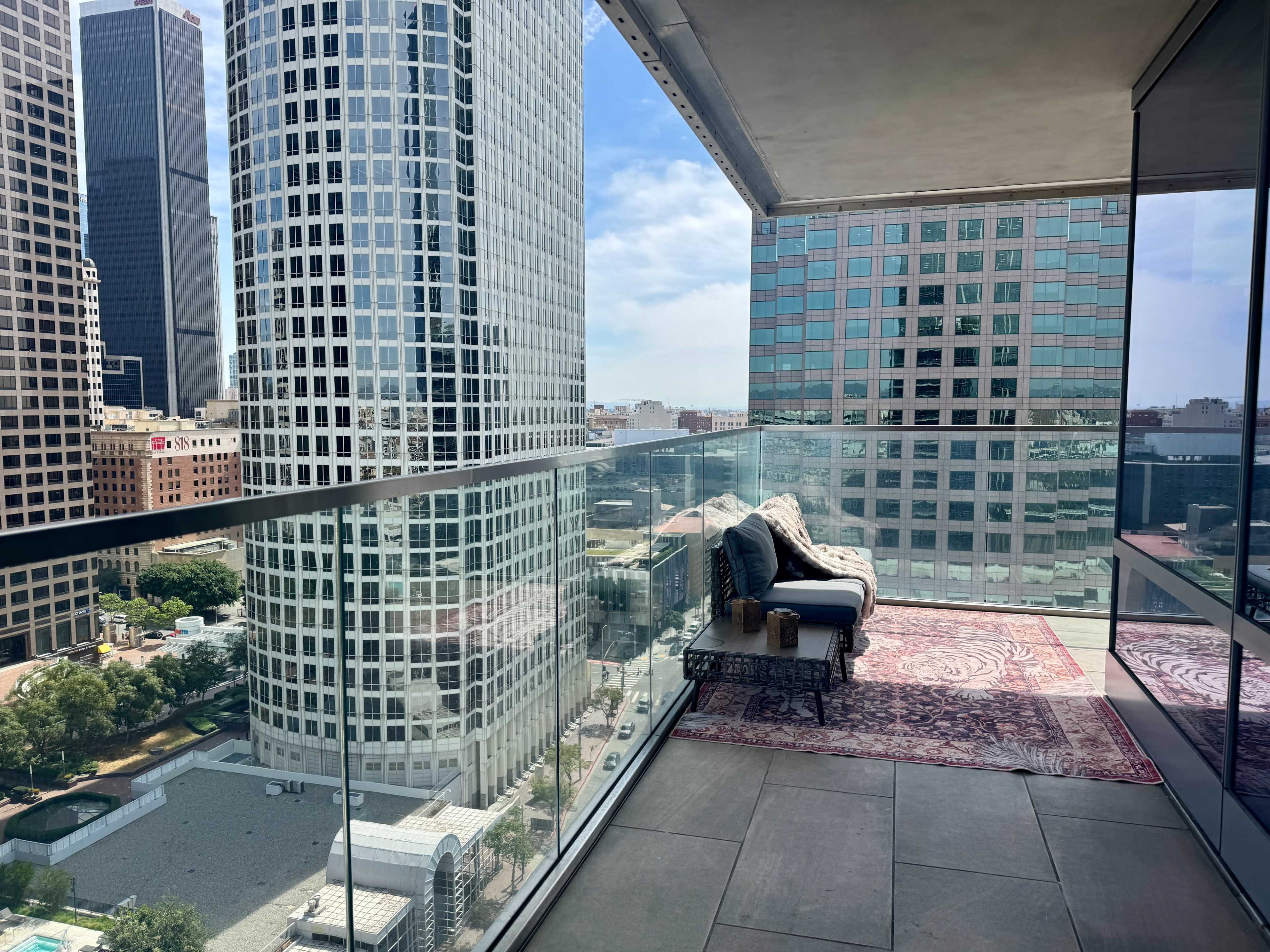 The image shows a glass balcony overlooking a cityscape with tall buildings, featuring a seating area and a patterned rug.