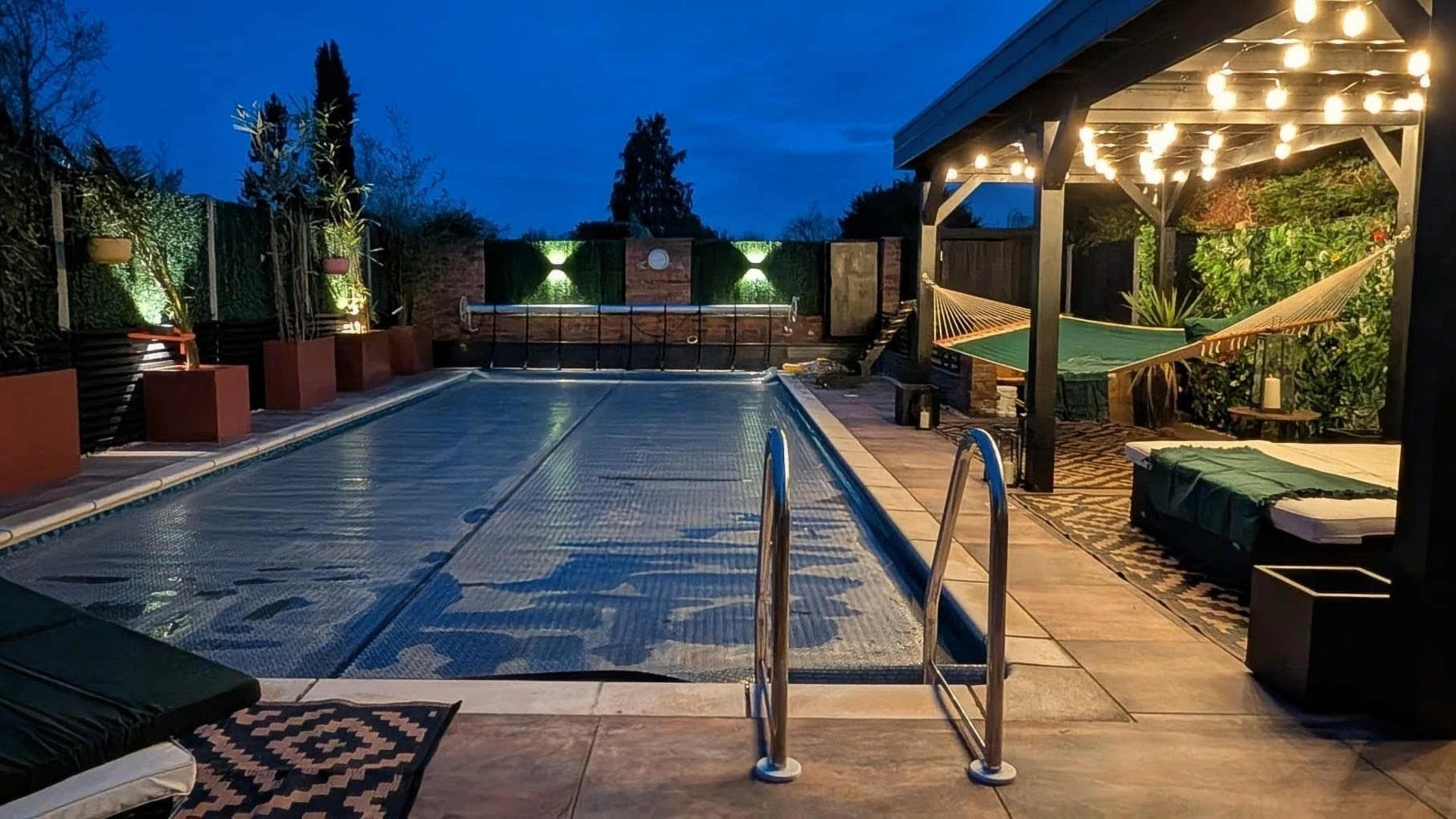 The scene features a swimming pool area at dusk, with illuminated hanging lights, a hammock, and surrounding greenery.