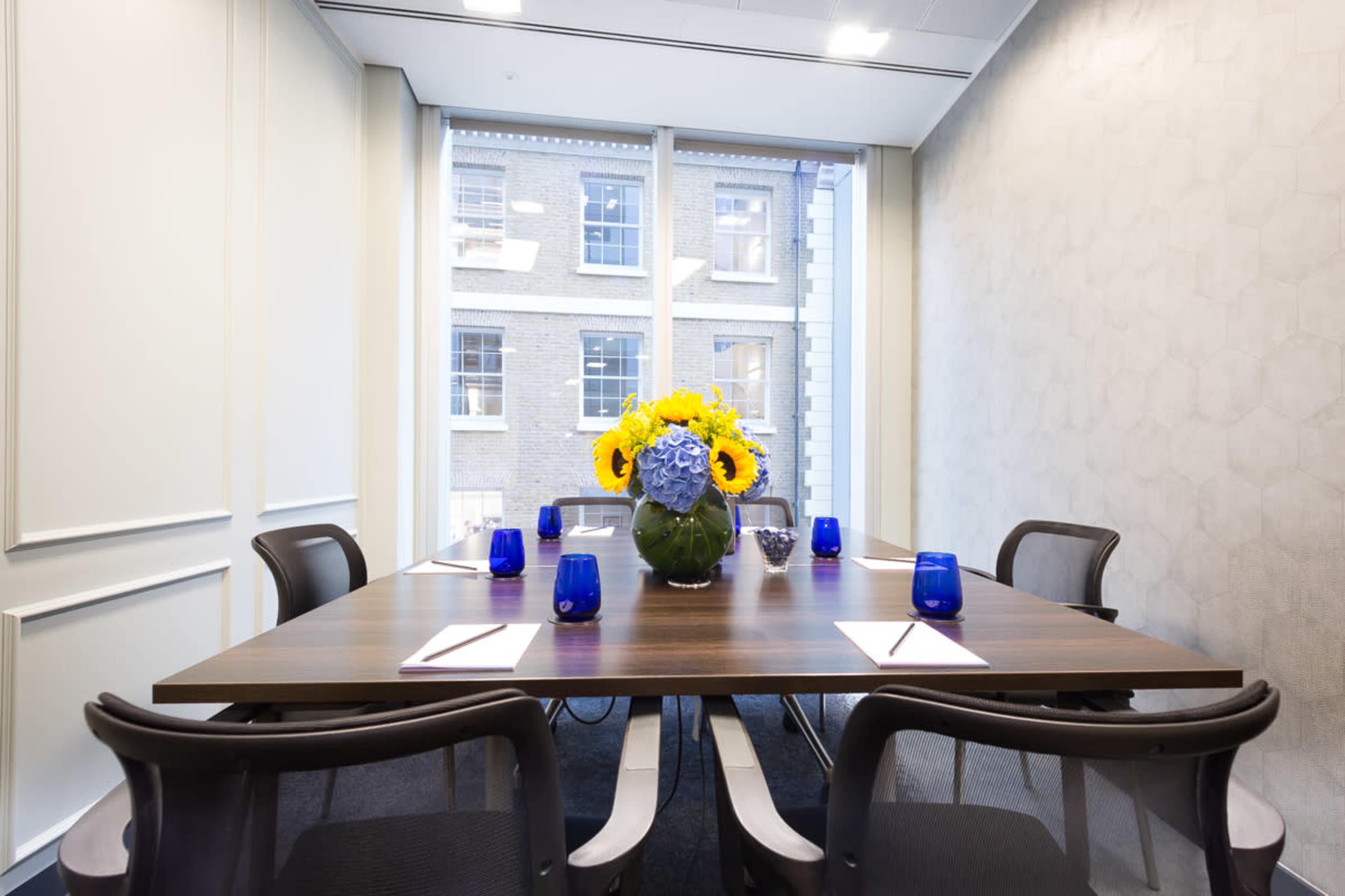 A meeting room features a wooden table set with notepads and blue glasses, centered around a vase of yellow flowers.