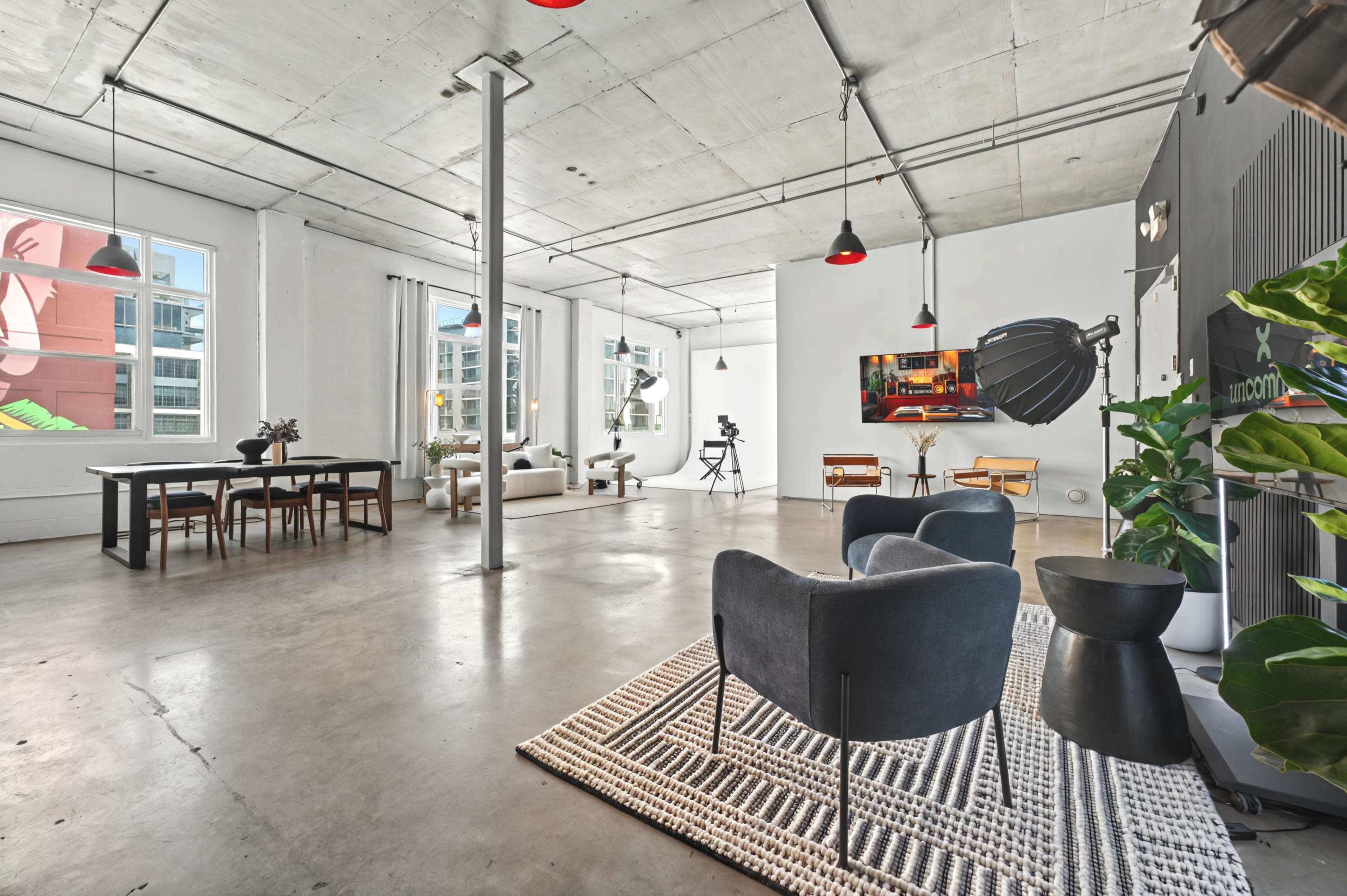 The image shows a spacious, modern loft with concrete floors, large windows, and a mix of dining and living areas furnished with various seating arrangements.
