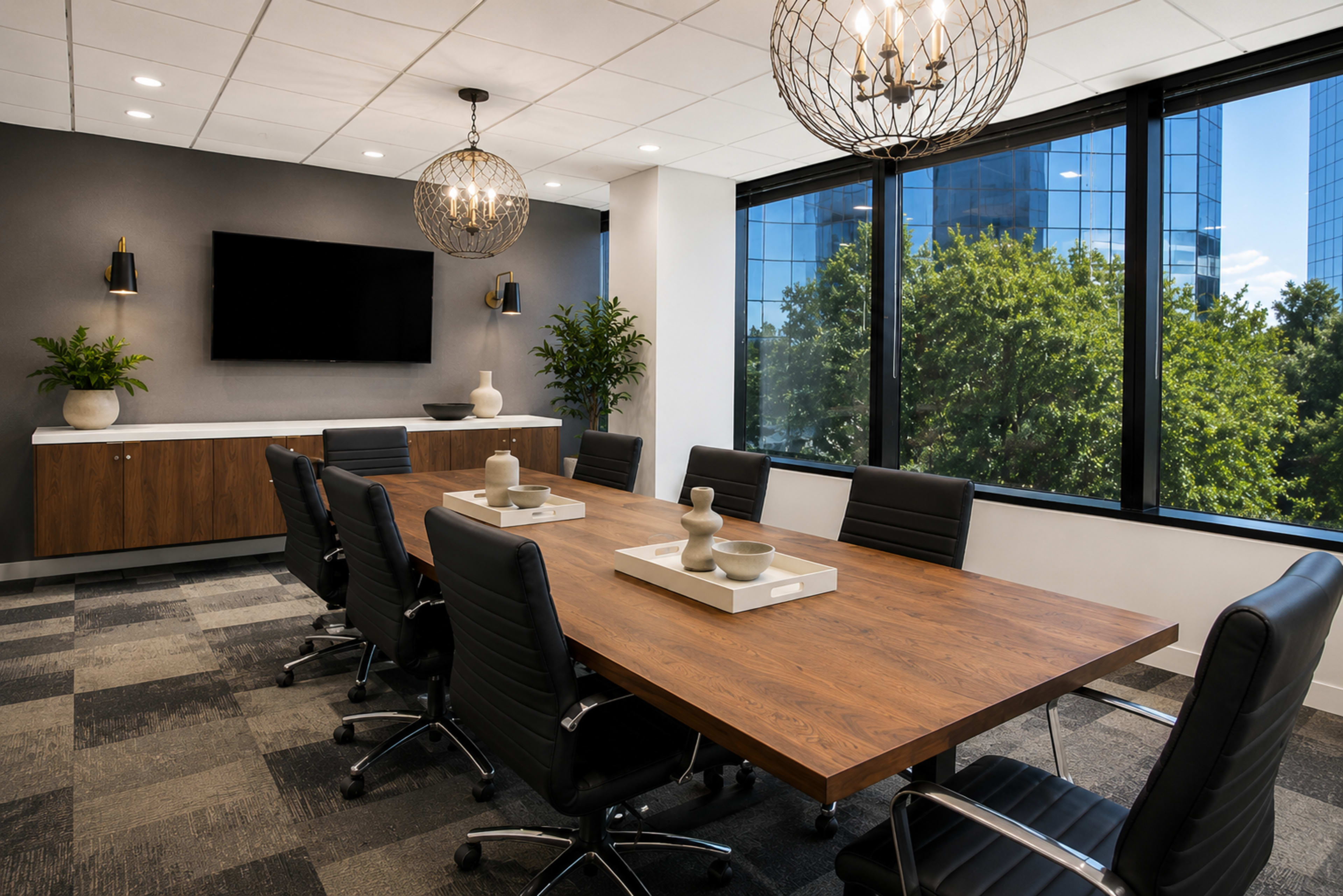 A modern conference room features a long wooden table surrounded by black leather chairs, with large windows overlooking trees and buildings outside.