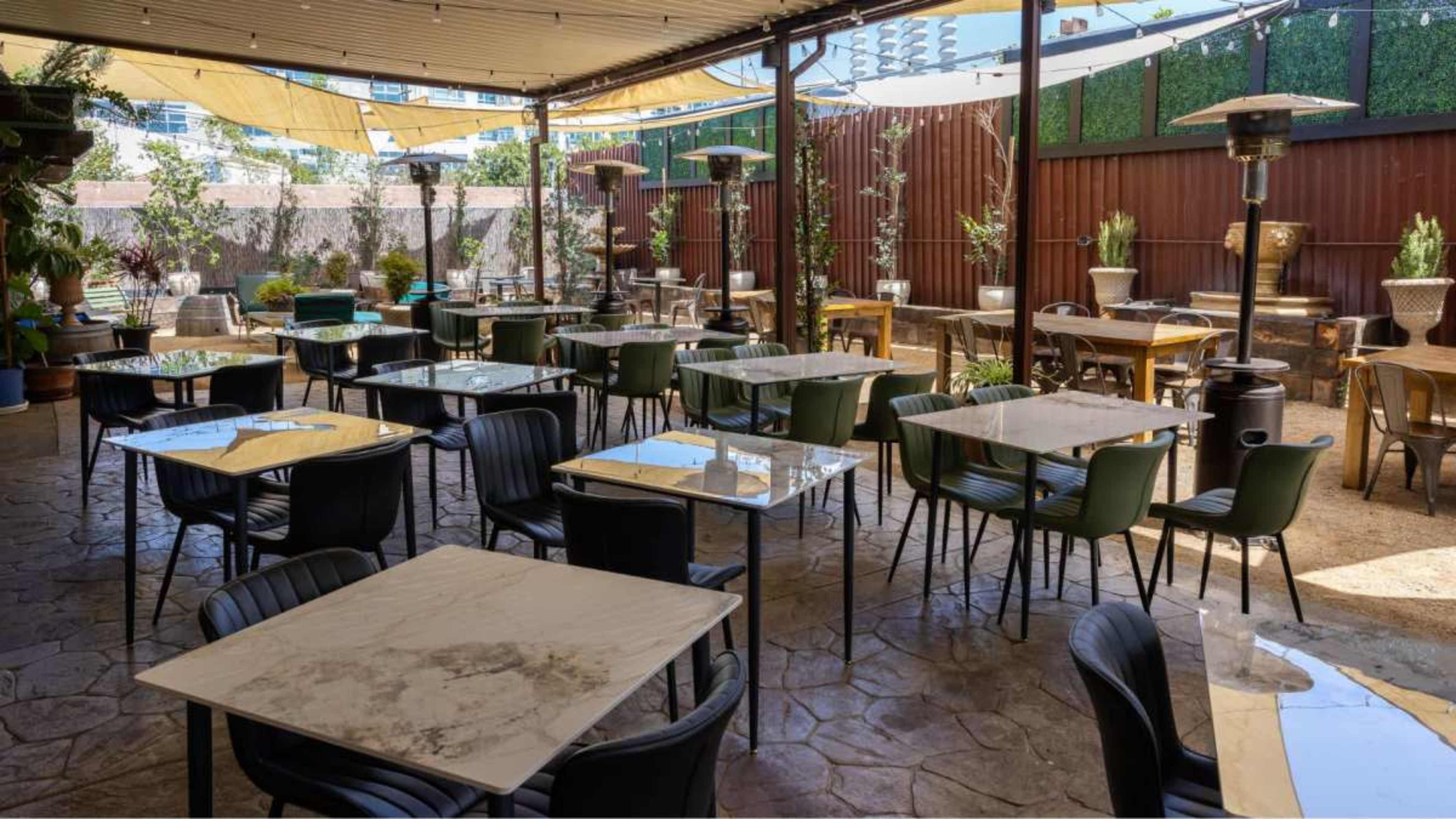The image shows an outdoor restaurant patio featuring rows of tables and chairs arranged under a shaded canopy.
