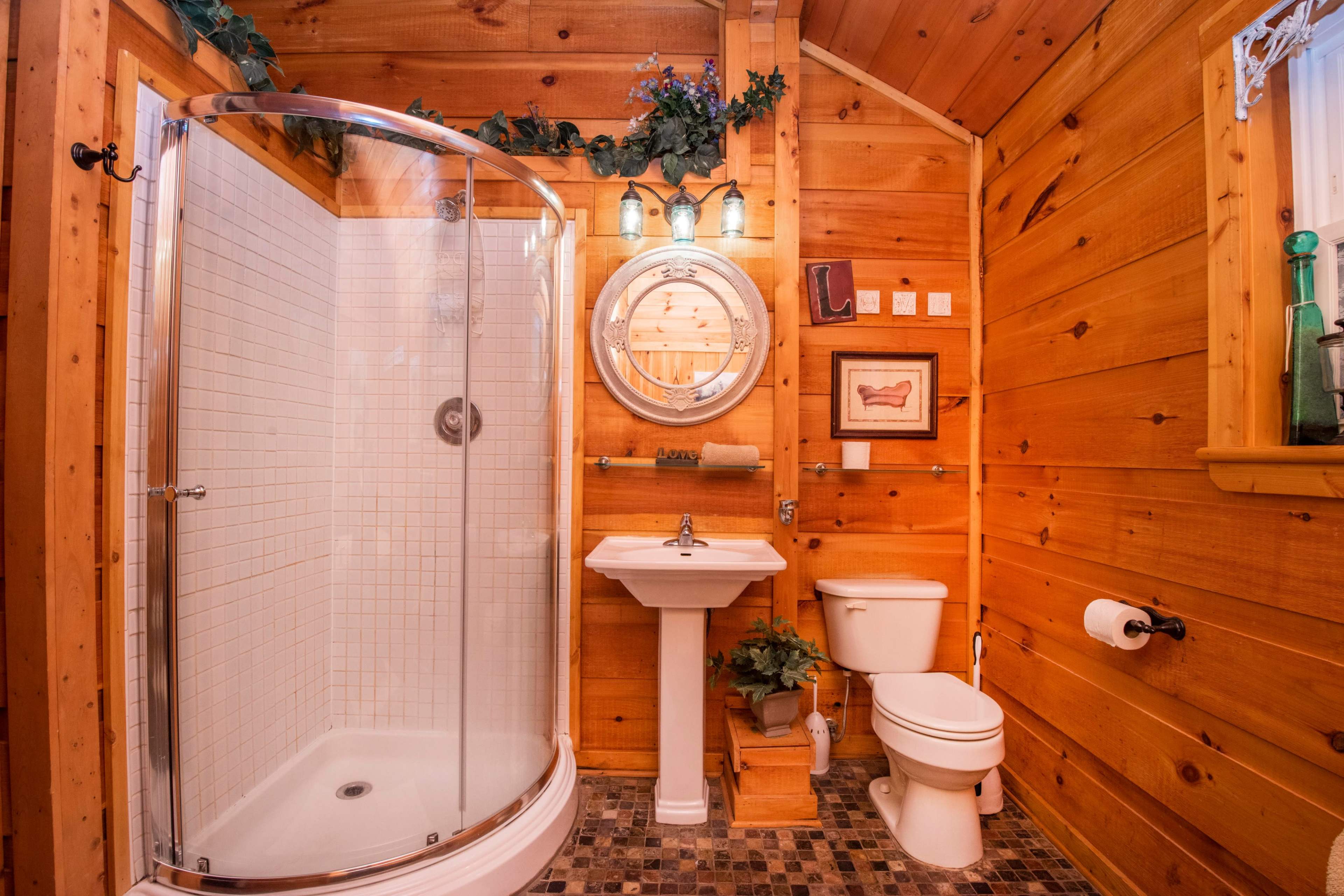 A wooden bathroom featuring a shower, sink, toilet, and décor elements such as plants and a circular mirror.