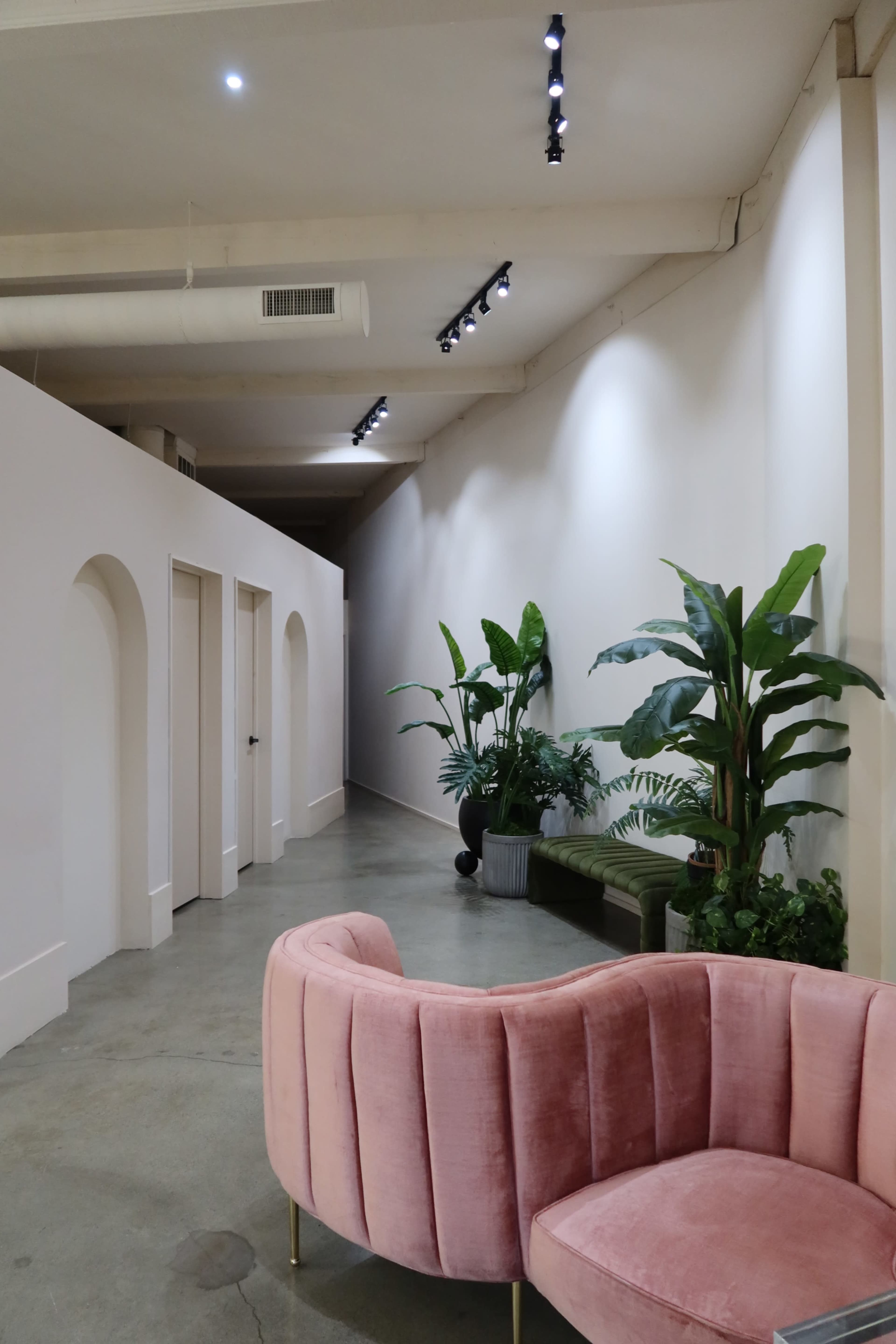 A narrow hallway features a pink velvet sofa, greenery in pots, and softly lit ceiling lights.