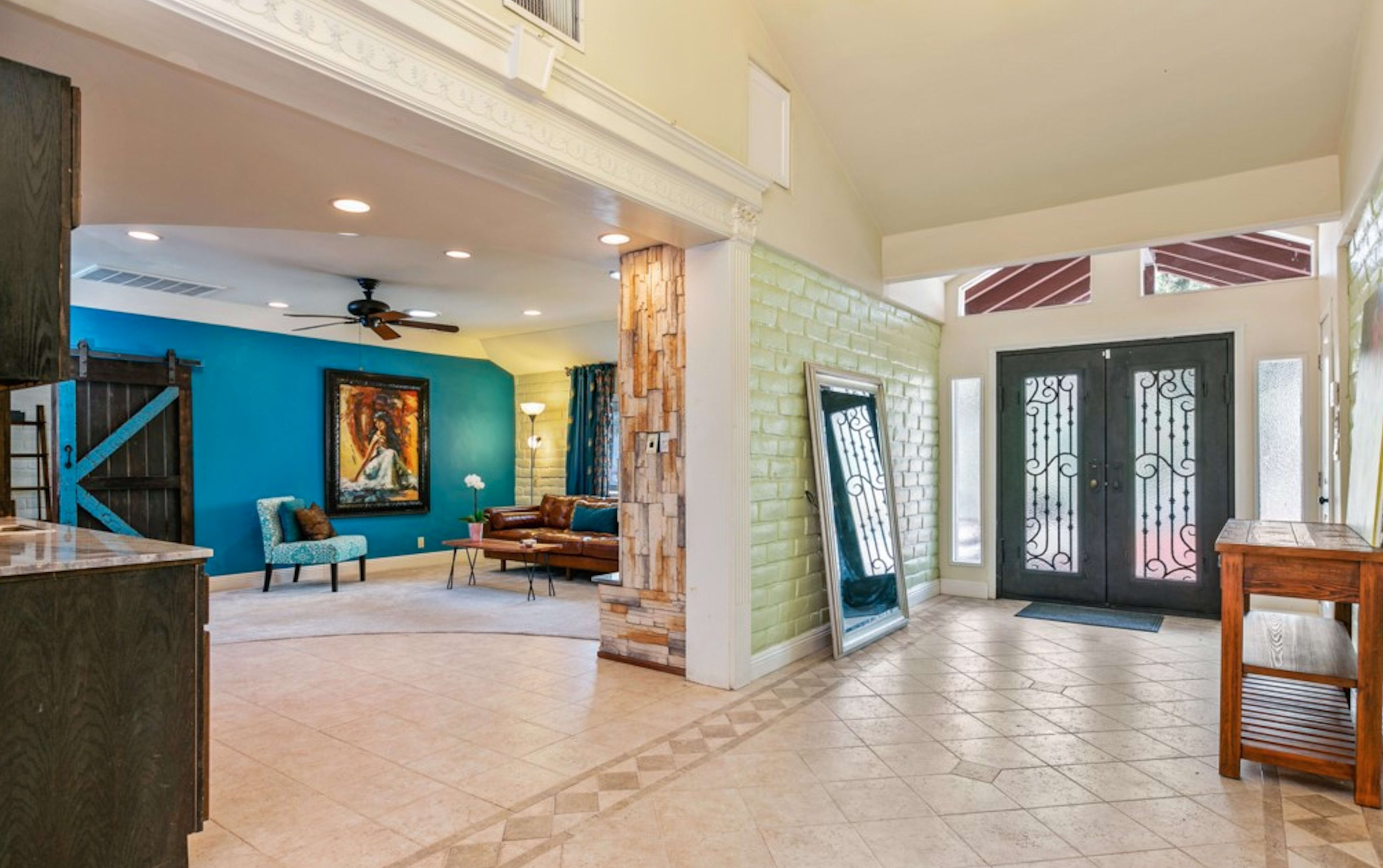 A spacious entryway featuring a tiled floor, a wooden table, two doors with decorative ironwork, and a living area with blue walls, a large artwork, and a wooden sliding door.