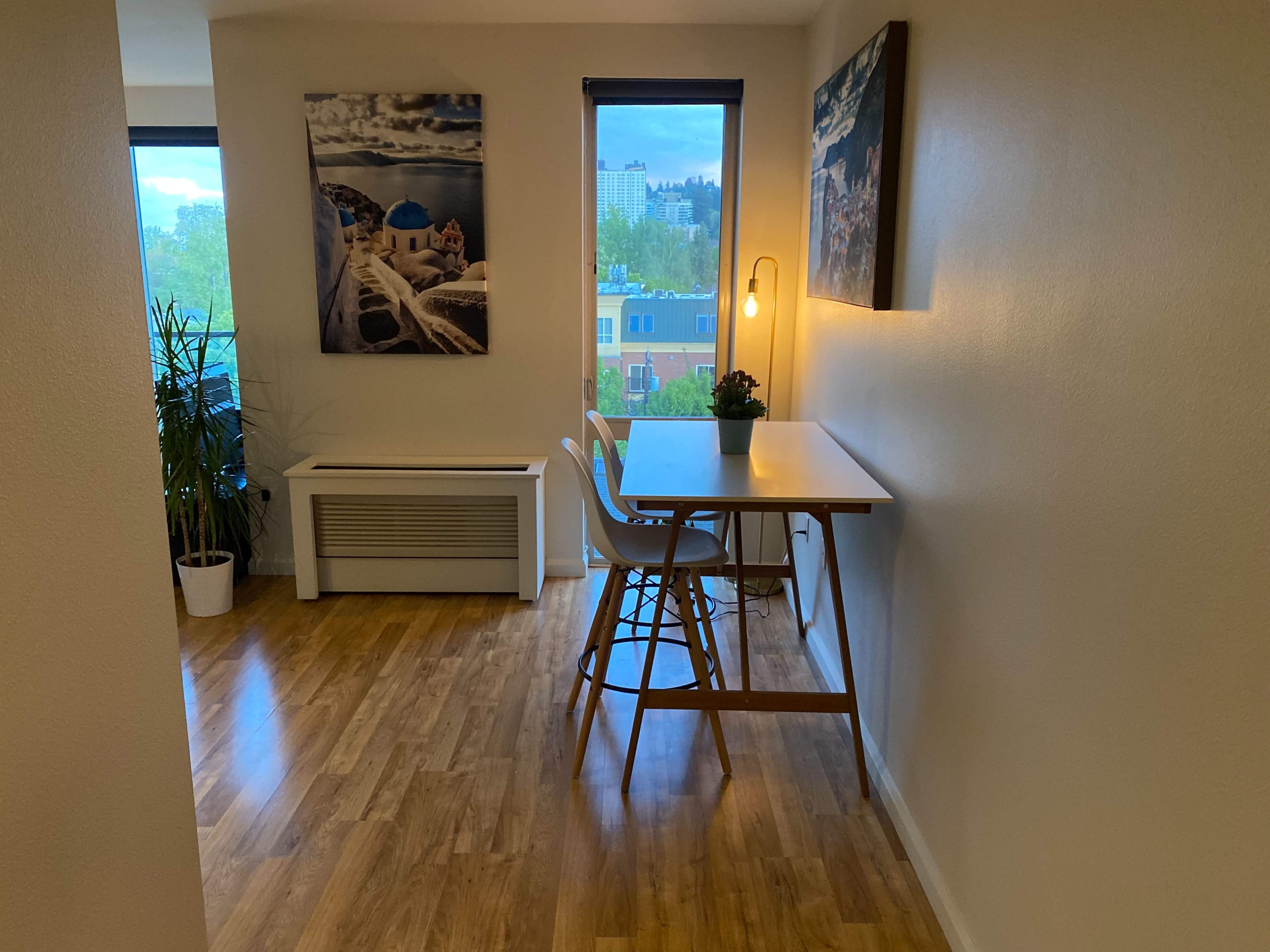 A small dining area features a table with two chairs, a potted plant, and a lamp, set against a backdrop of a window showcasing a city view.