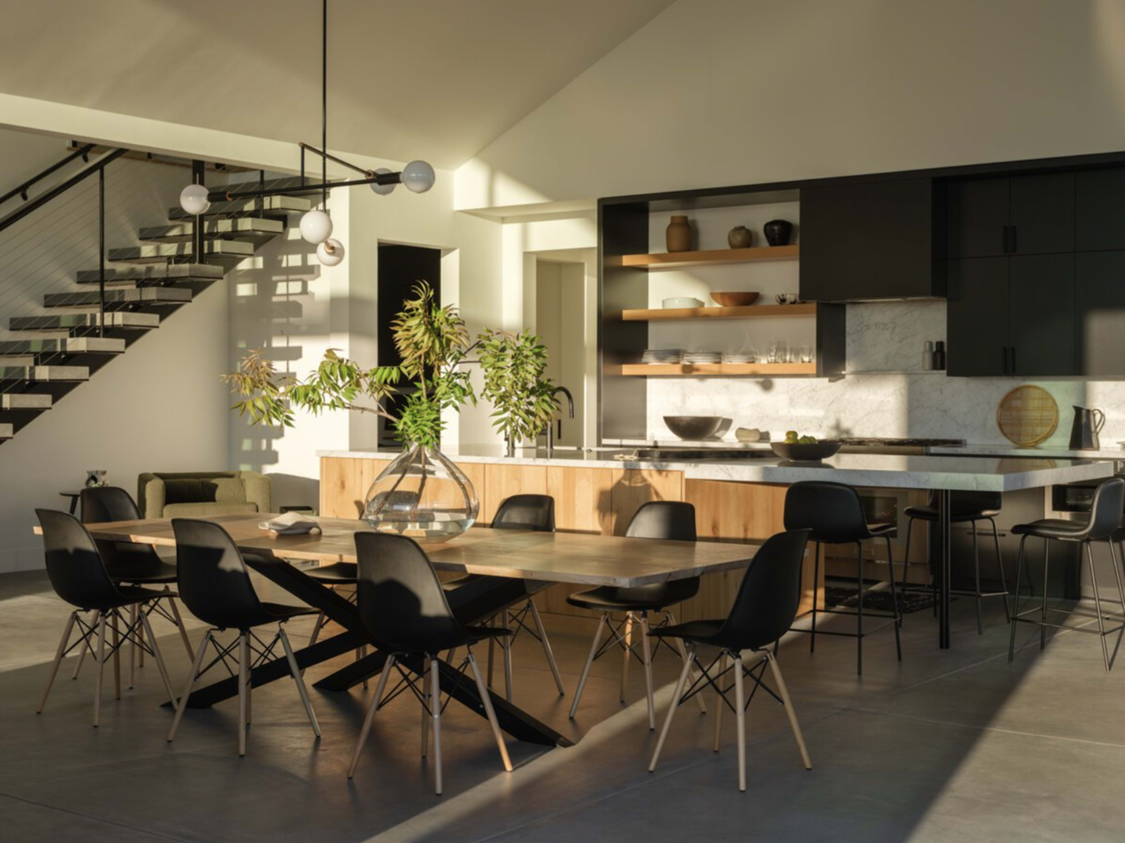 A spacious, modern dining area features a large table surrounded by black chairs, with a staircase and a kitchen area visible in the background.