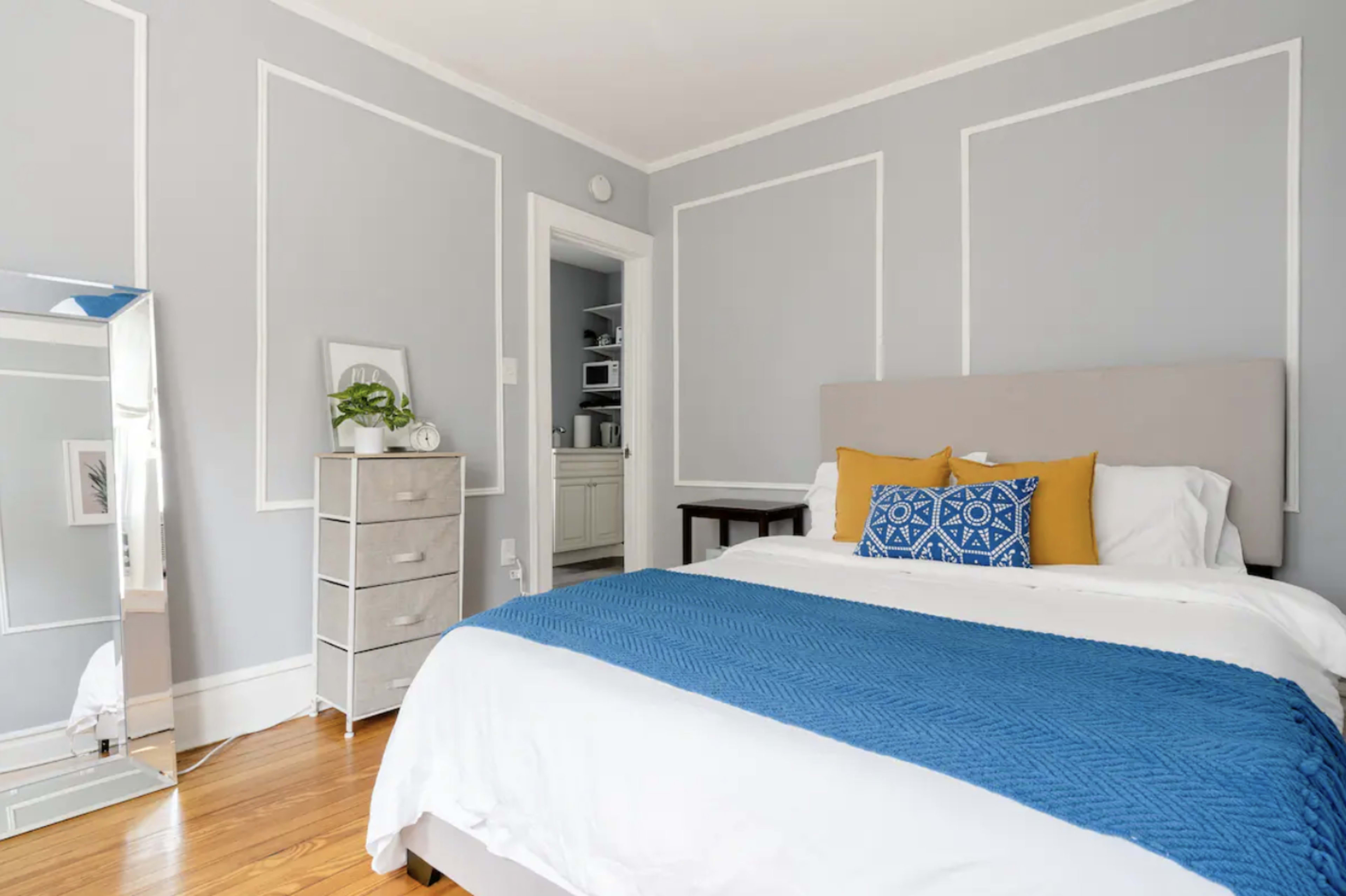 A neatly arranged bedroom features a bed with blue and yellow accents, a mirrored dresser, and light gray walls with white trim.