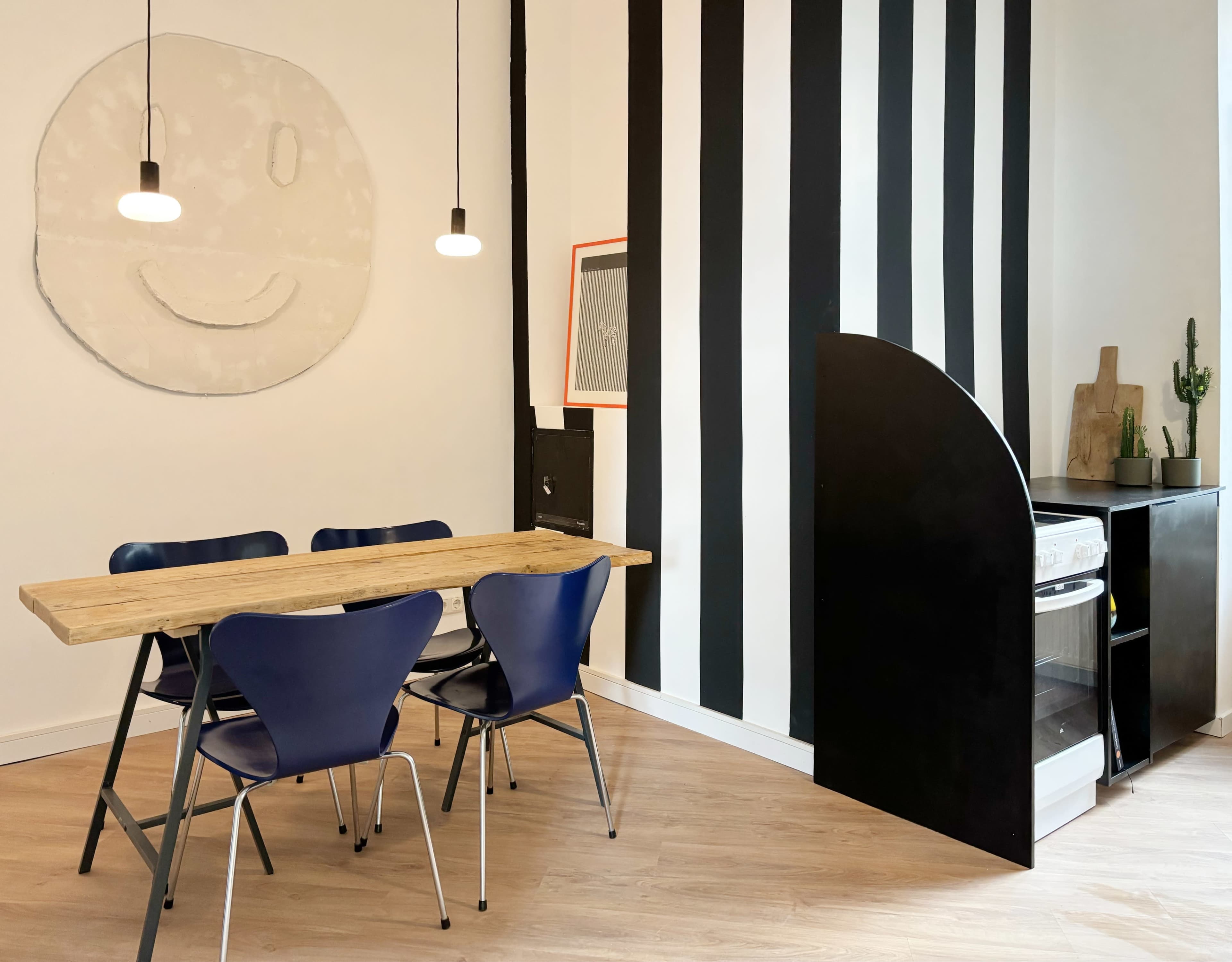 A minimalist dining area features a wooden table surrounded by blue chairs, with a black and white striped wall and a small refrigerator.