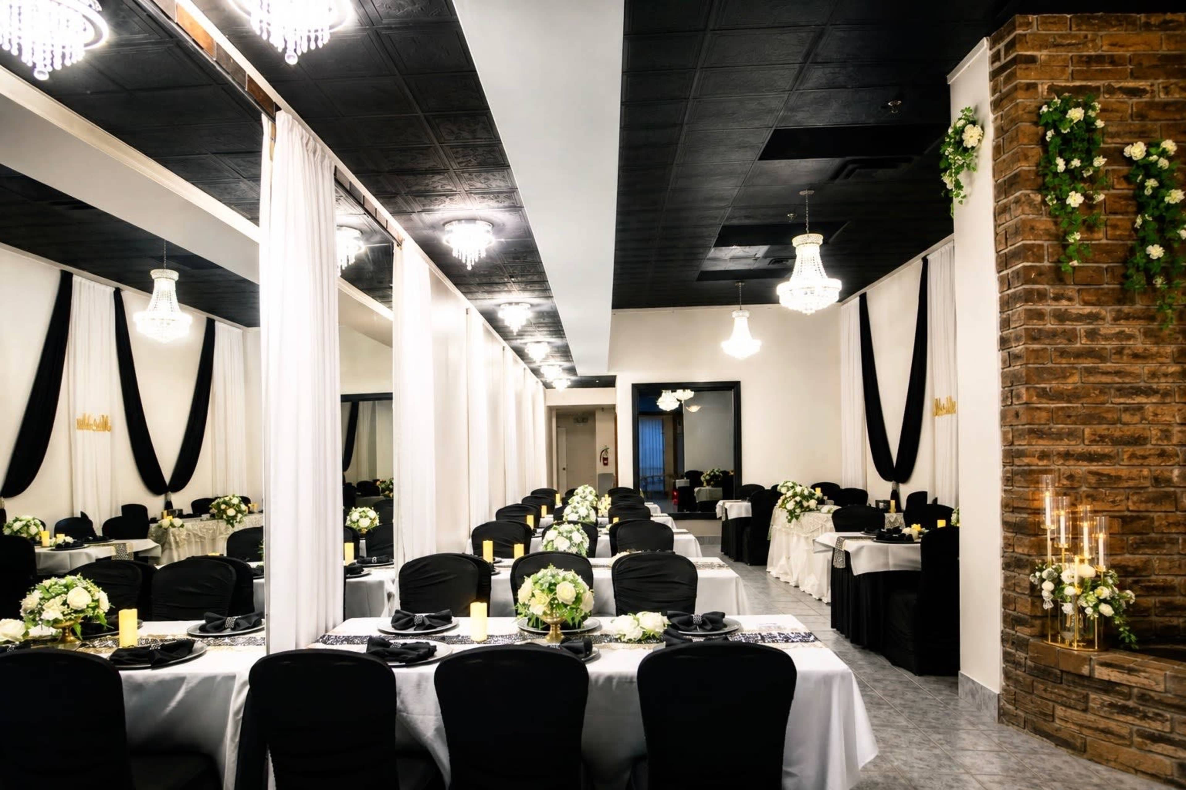 The image shows a decorated event space with tables set for a formal gathering, featuring white tablecloths, black chairs, floral centerpieces, and ambient lighting from chandeliers.