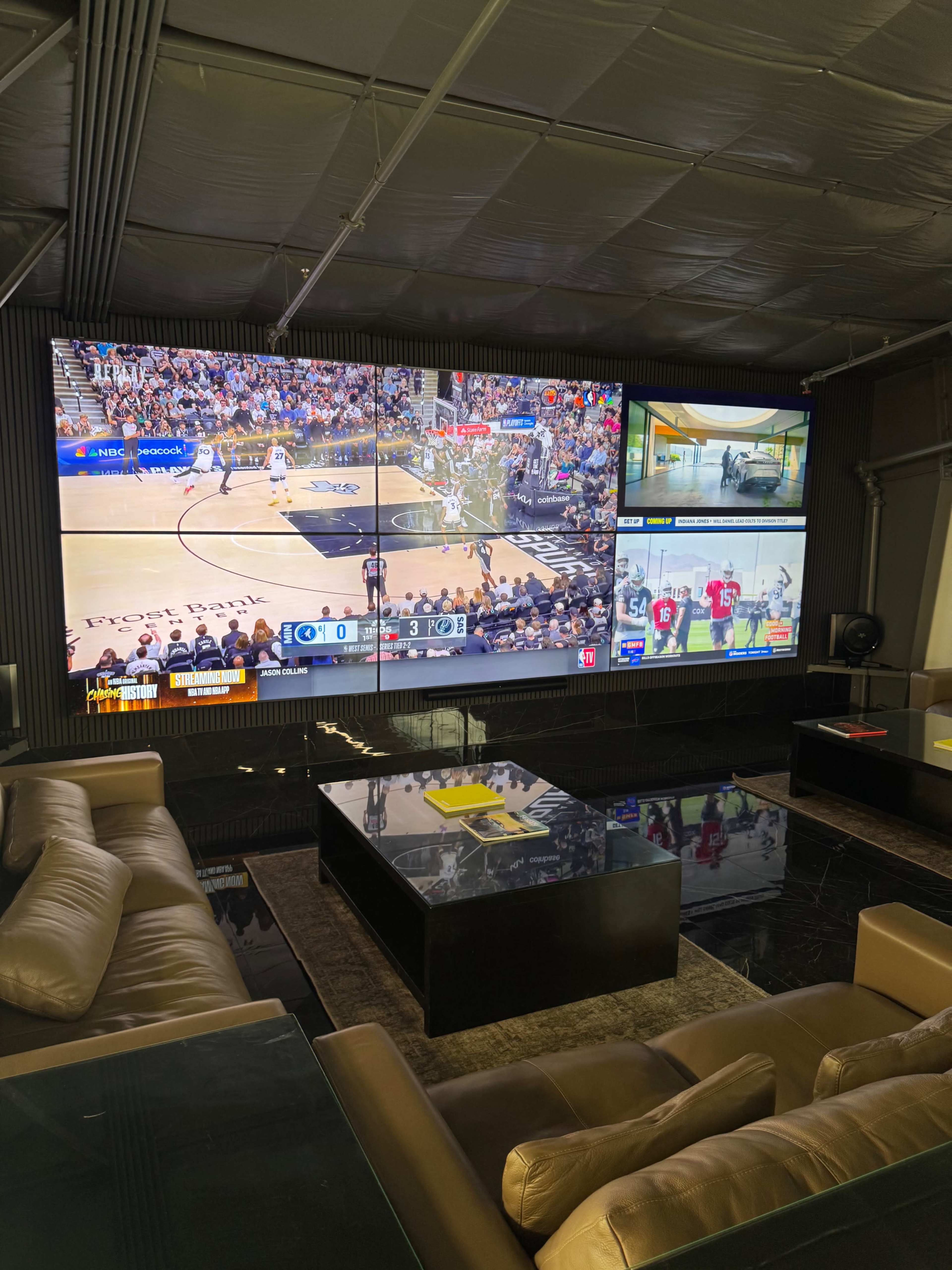 The image shows a sports lounge with multiple large screens displaying different games, featuring a comfortable seating area with a dark table in the center.