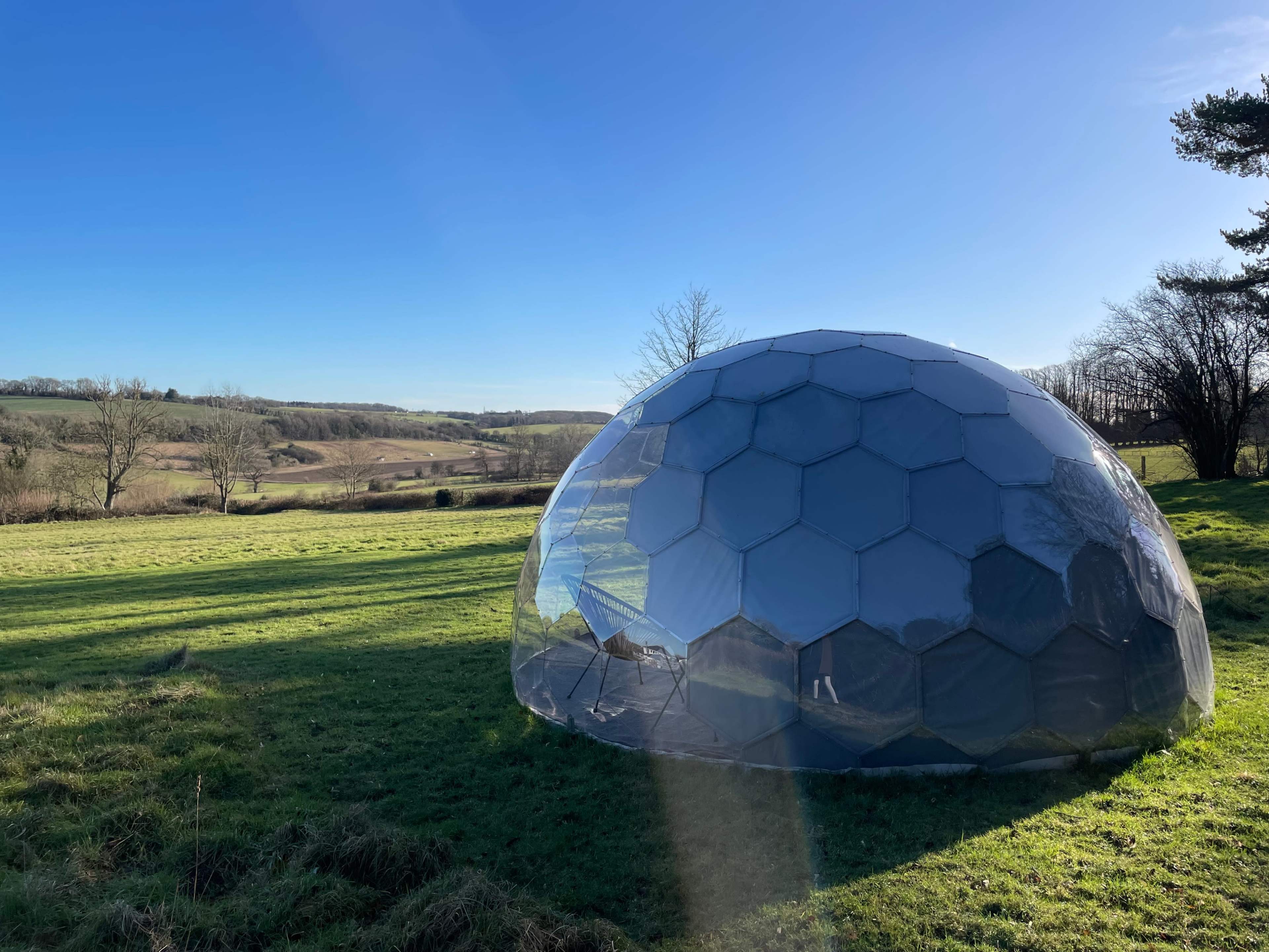 A transparent, geodesic dome structure is situated on a grassy hillside overlooking a valley under a clear blue sky.