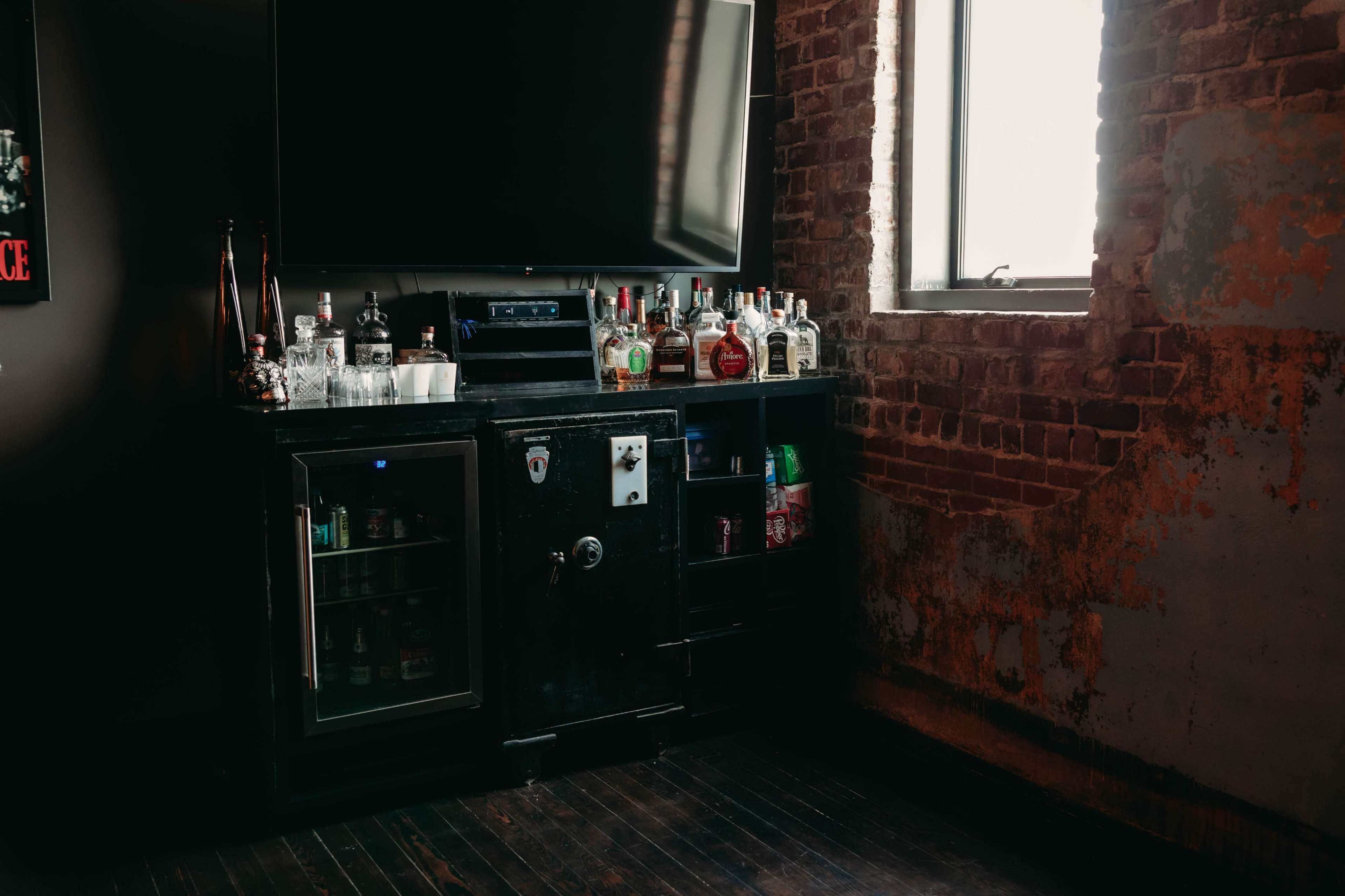 A home bar features a television mounted on the wall, surrounded by various bottles of liquor and a mini-fridge on a wooden floor beneath a window.