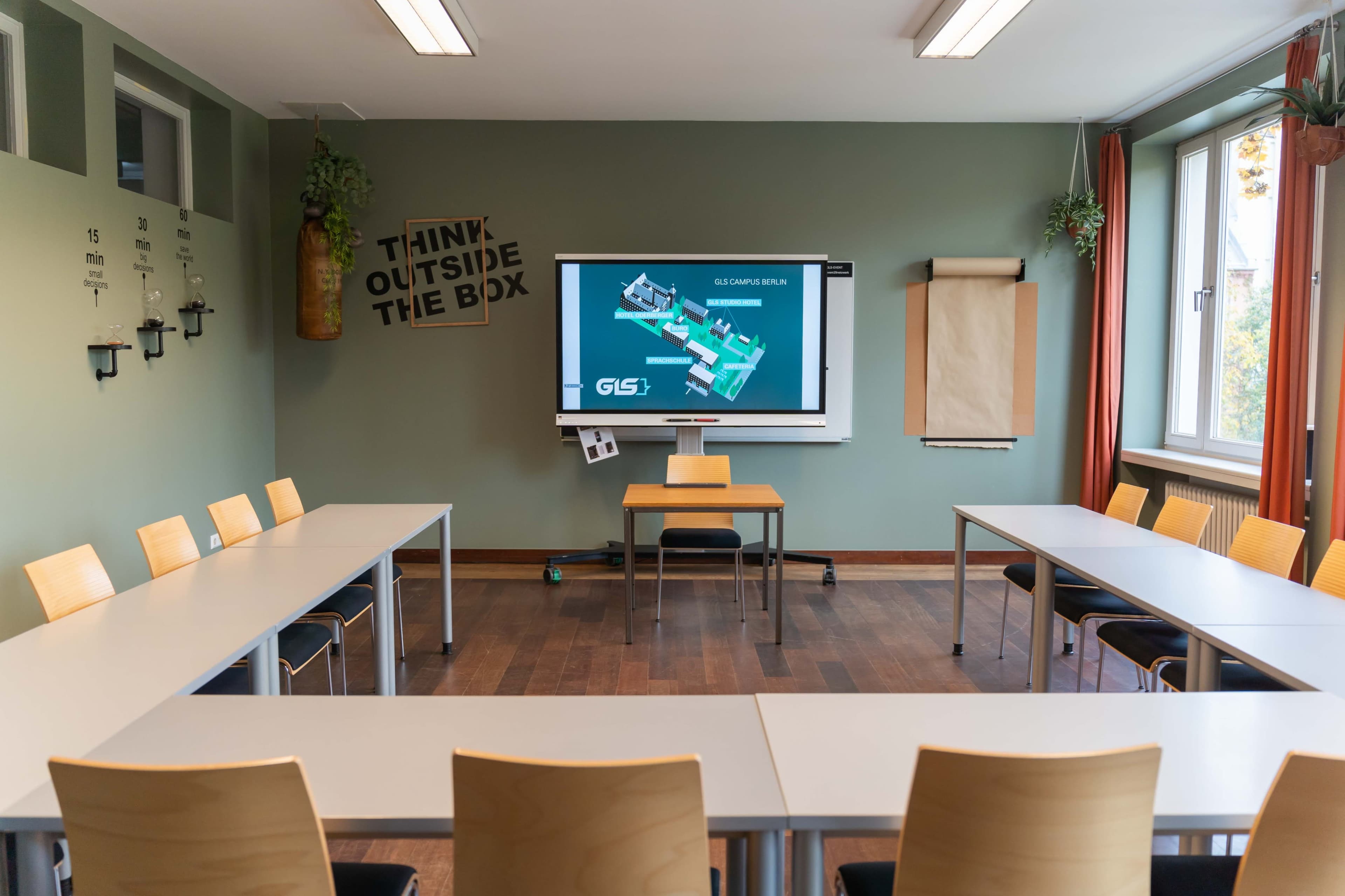 A classroom is arranged with tables in a U-shape, featuring a projector screen displaying a presentation, and walls adorned with minimal decorations.