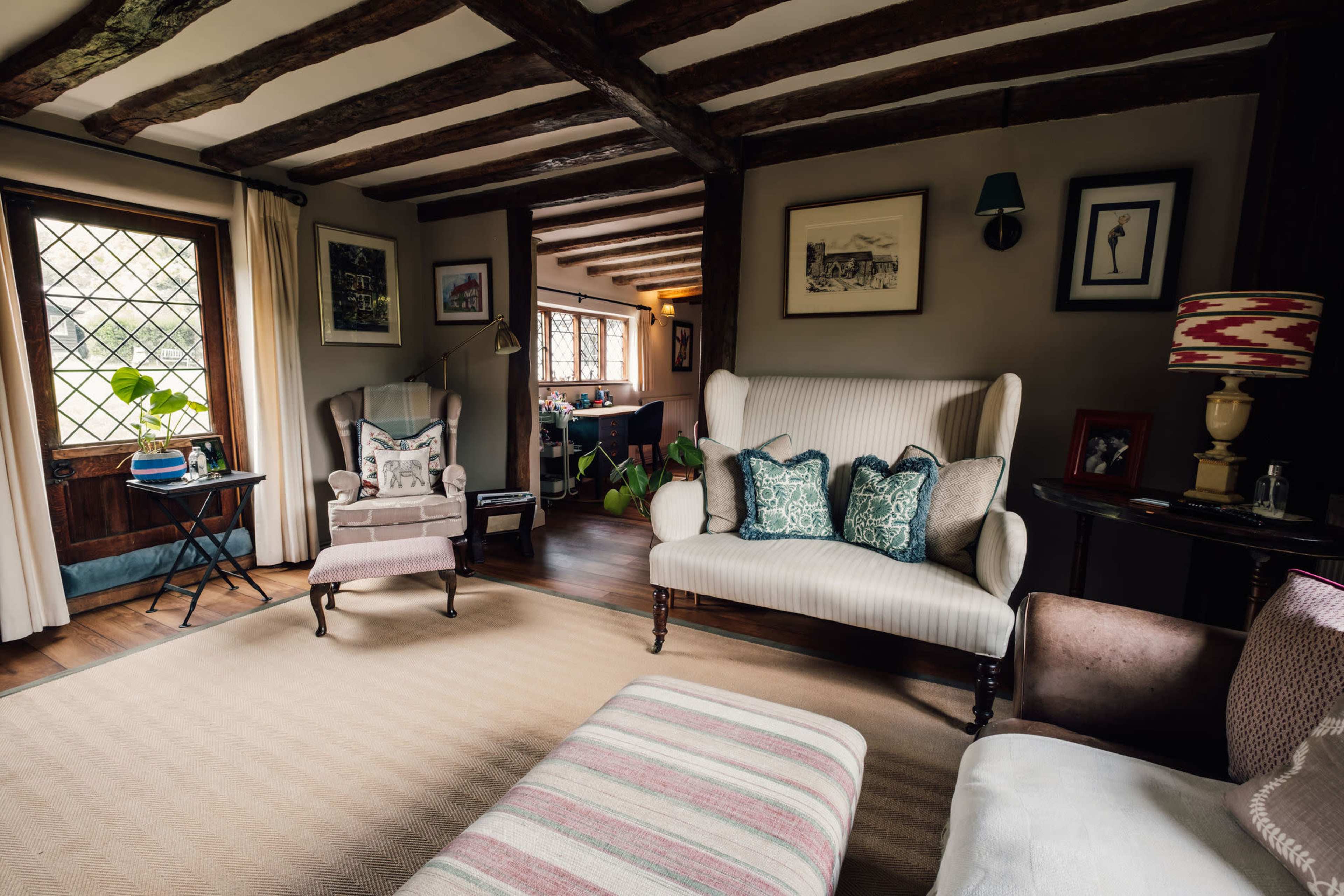 The image shows a cozy living room featuring two upholstered armchairs, a sofa with decorative pillows, wooden beams on the ceiling, and a window that lets in natural light.