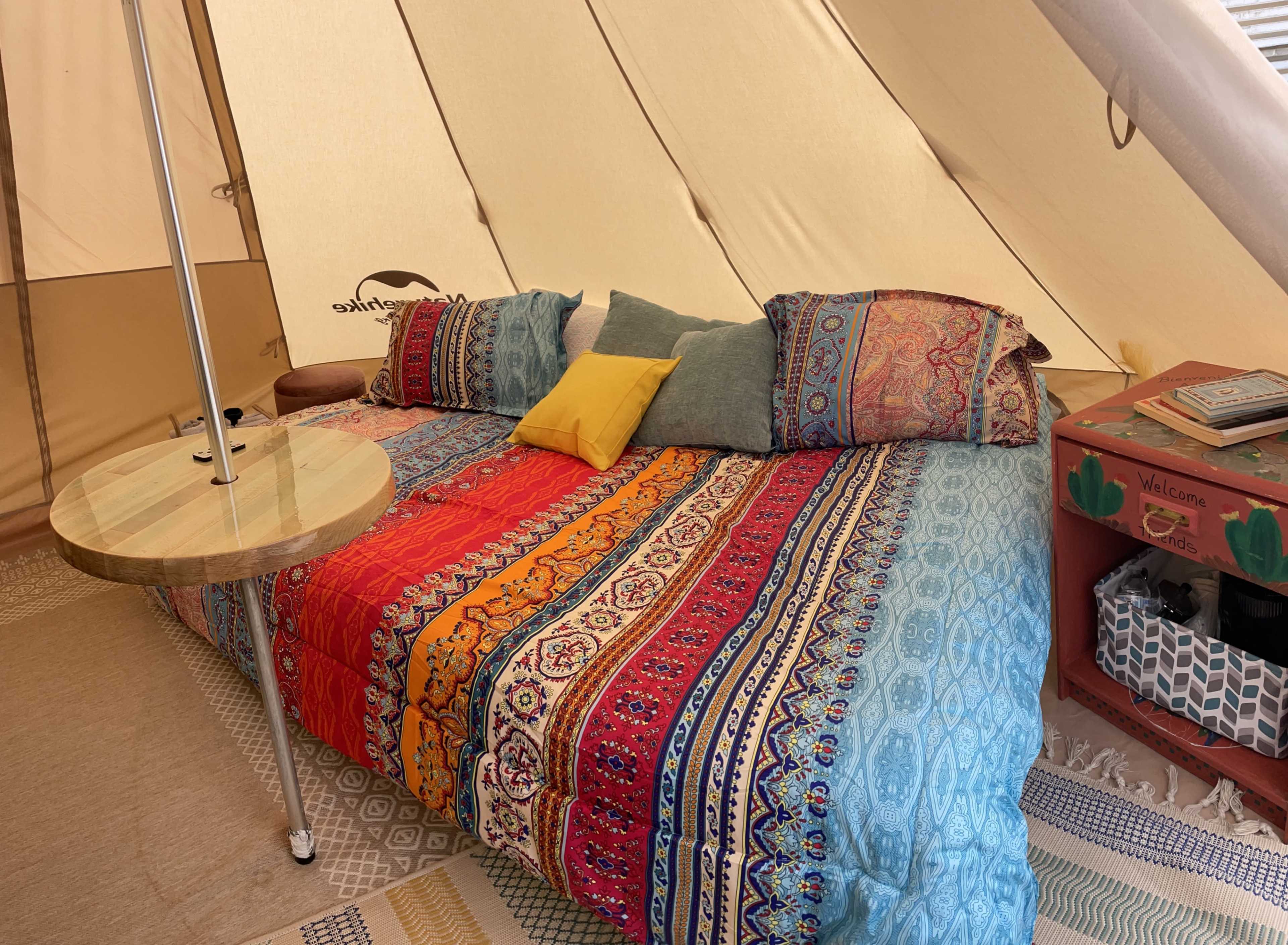The image shows a neatly arranged bed inside a canvas tent with colorful patterned bedding and cushions, alongside a small side table and a red storage unit.