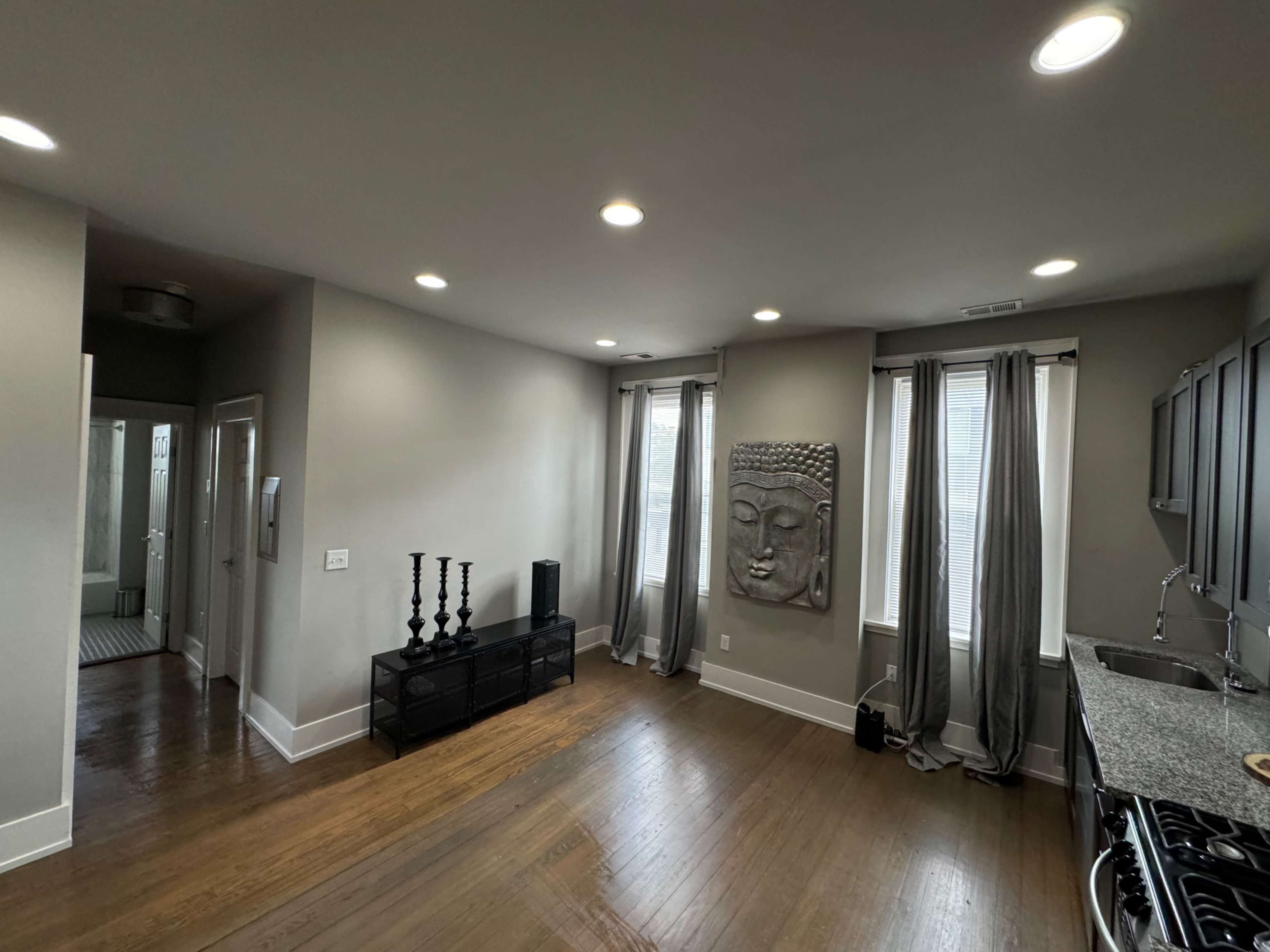 A spacious living area featuring a Buddha face wall decoration, sleek cabinets, and large windows with gray curtains.