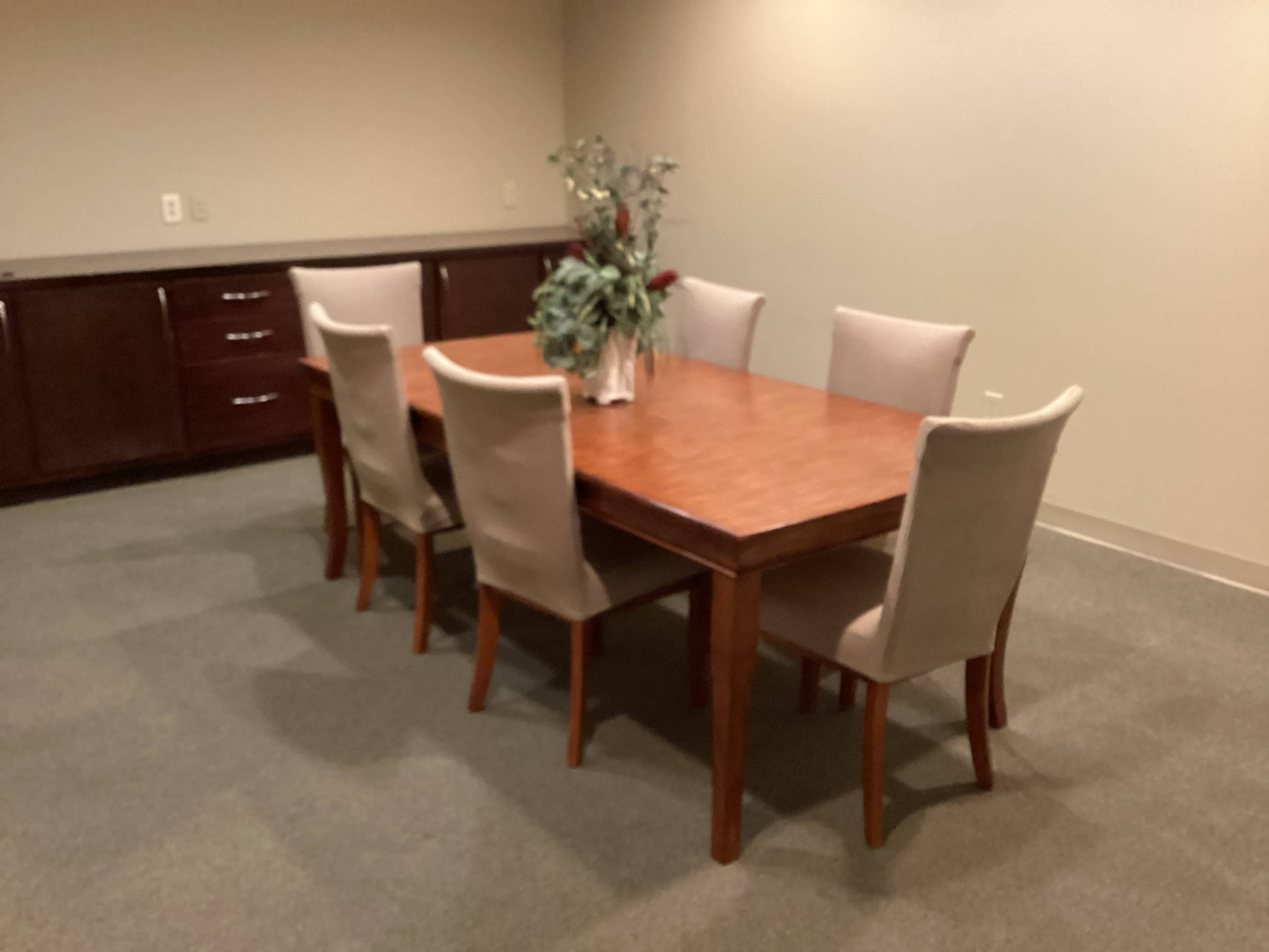 A wooden dining table is set in a room surrounded by six upholstered chairs, with a vase of flowers in the center.