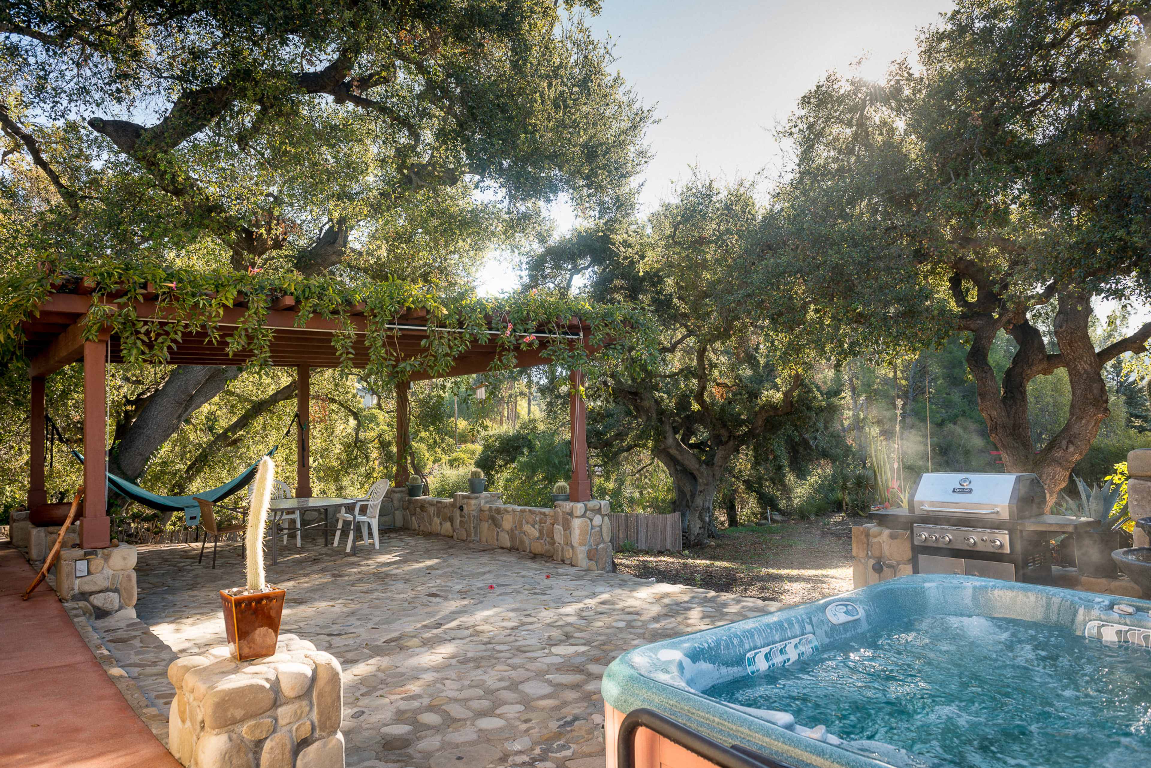 The image shows a patio area with a hot tub, a barbecue grill, a stone floor, and a hammock hanging under a wooden pergola surrounded by trees.