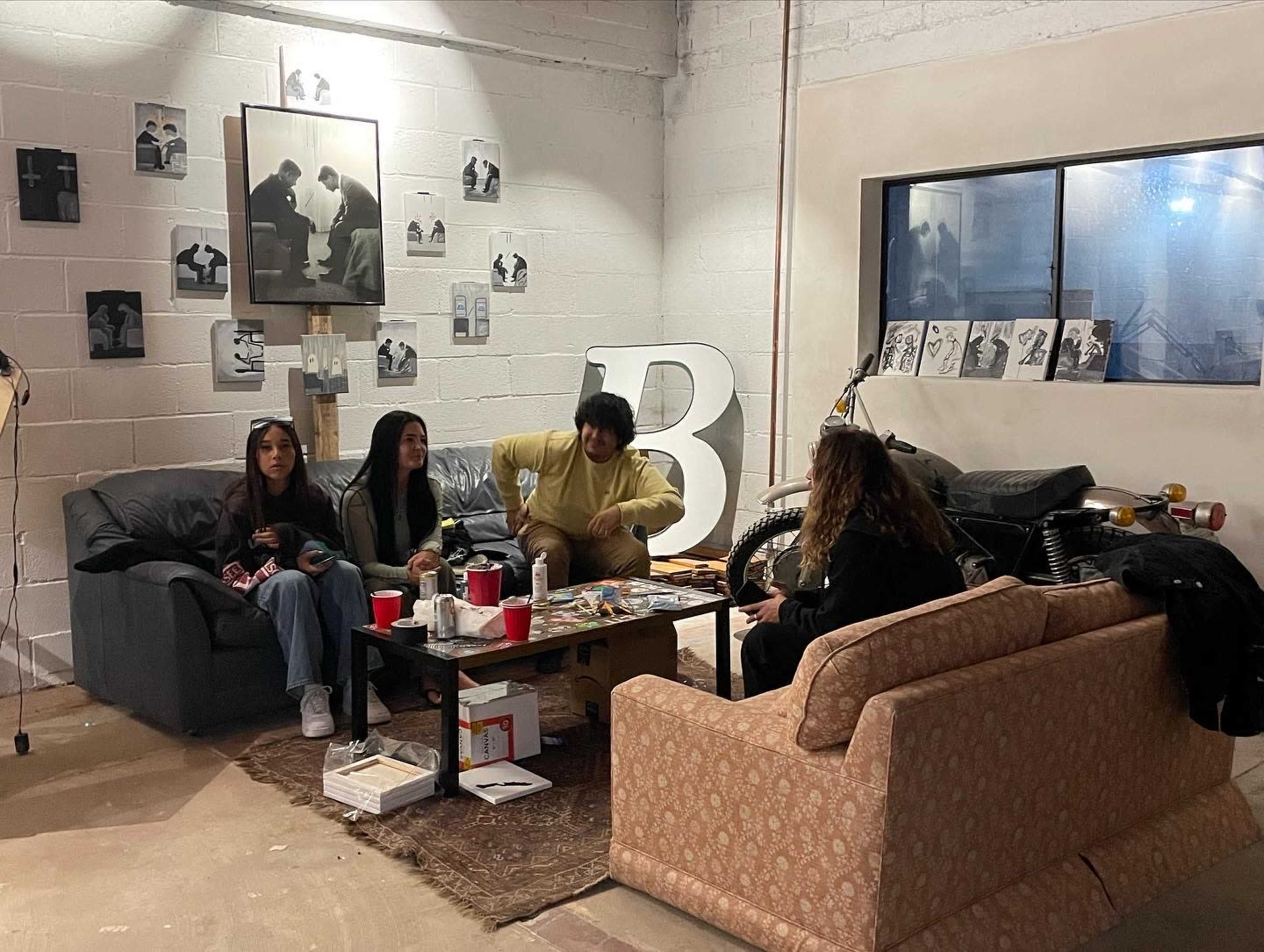 Four people are seated in a casual living space with a coffee table, surrounded by artwork on the walls and an old motorcycle in the background.