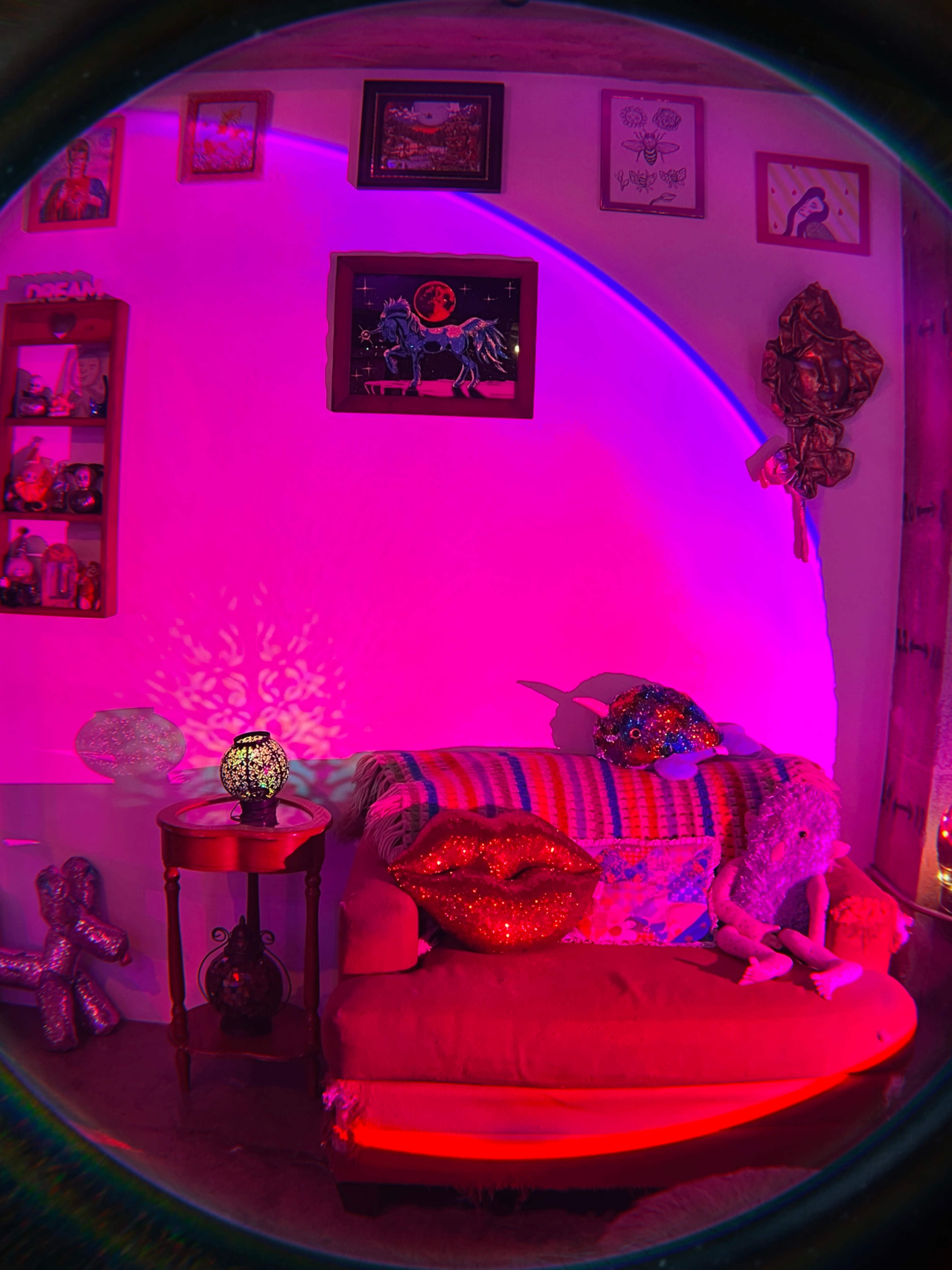 The image shows a vibrantly lit corner of a room featuring a red sofa adorned with decorative pillows, a small table with a lamp, and various wall decorations.