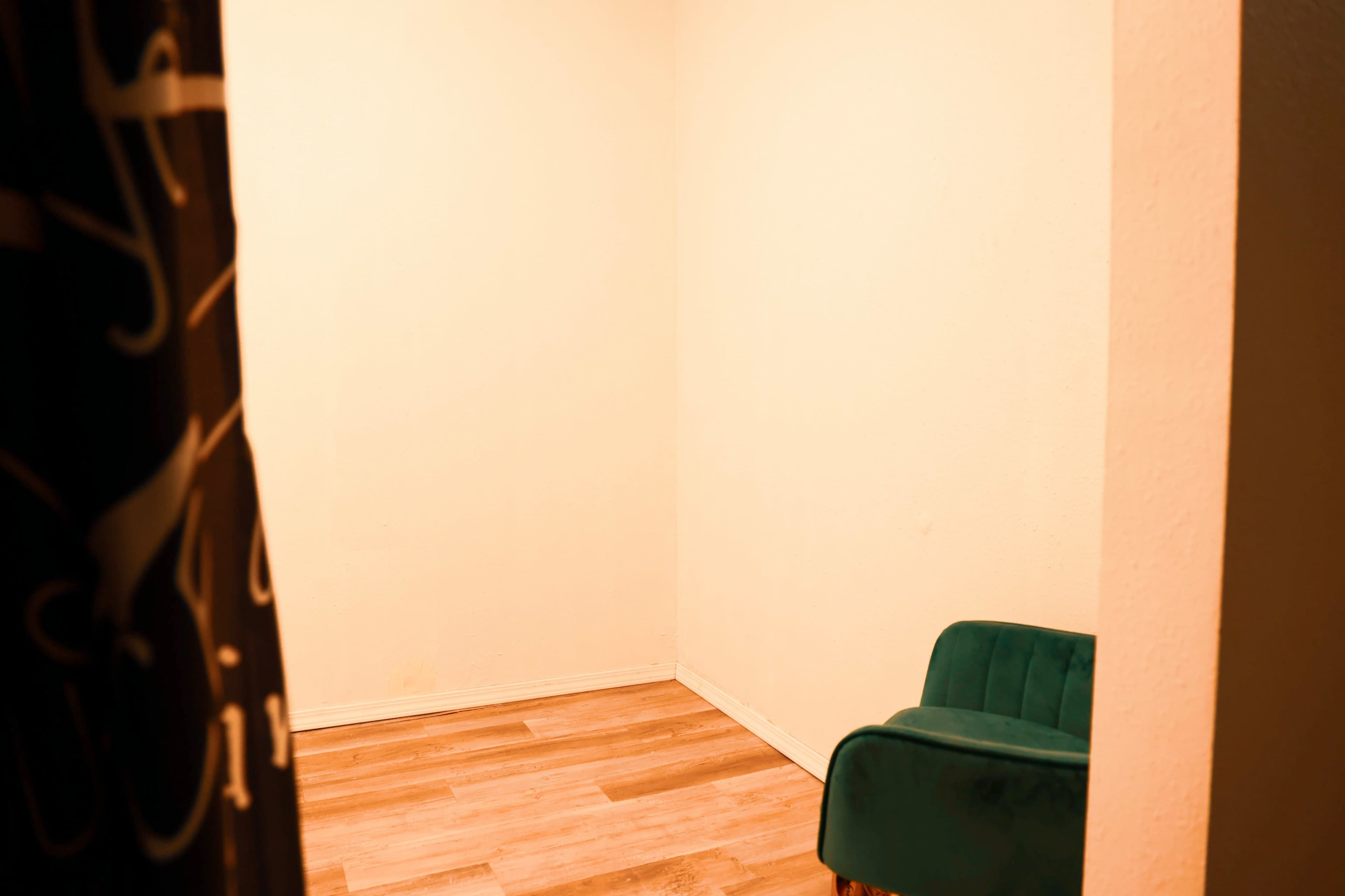A small, empty room with light-colored walls and a green chair positioned in one corner on wooden flooring.