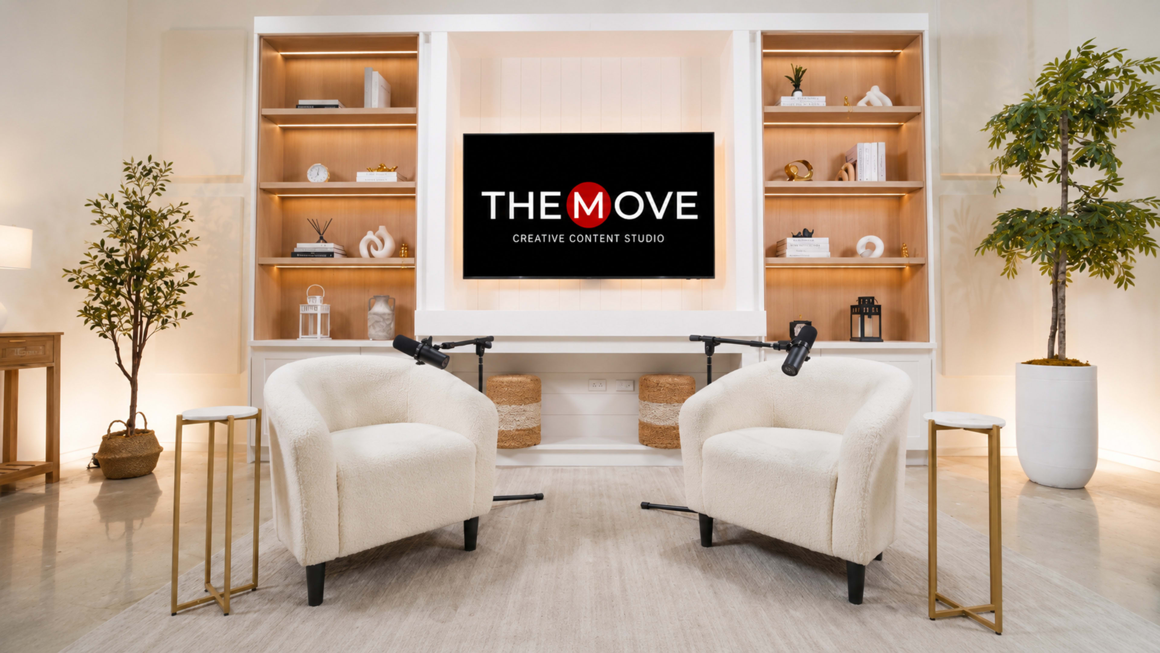 The image shows a modern creative content studio setup with two white armchairs, microphones positioned on stands, a television screen displaying "THE MOVE CREATIVE CONTENT STUDIO," and decorative shelves in the background.