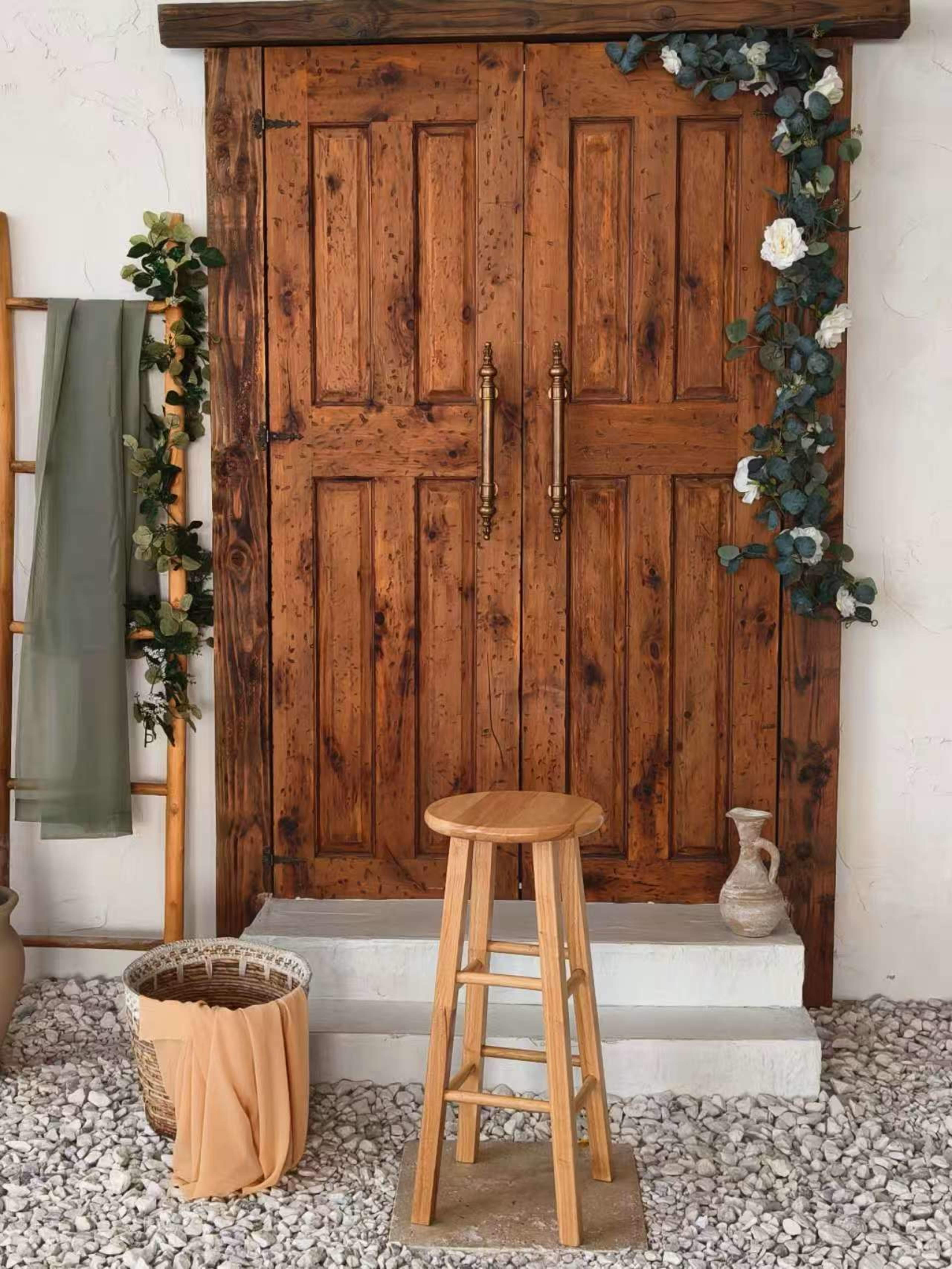The image shows a rustic wooden door framed by greenery and a stool in front, set against a backdrop of smooth pebbles.