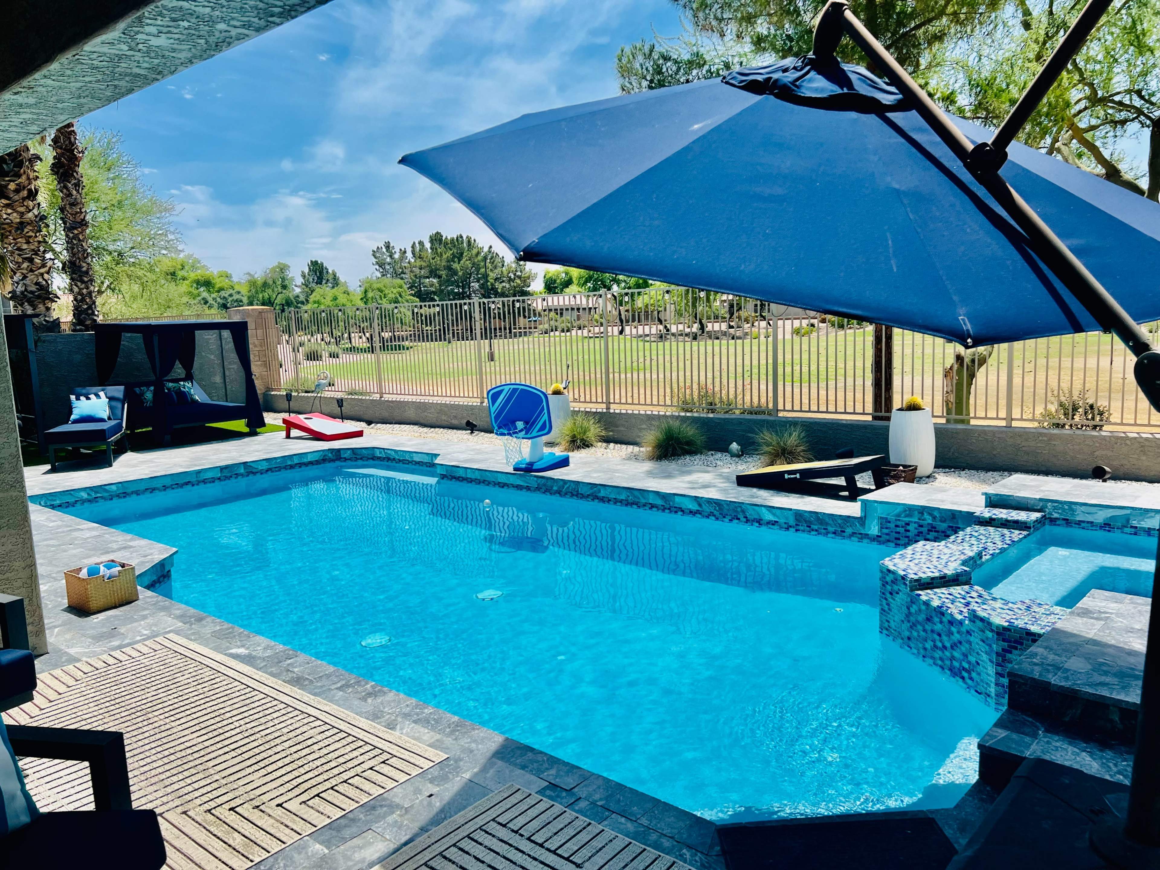 A blue swimming pool surrounded by lounge chairs and an umbrella, with a landscaped view in the background.