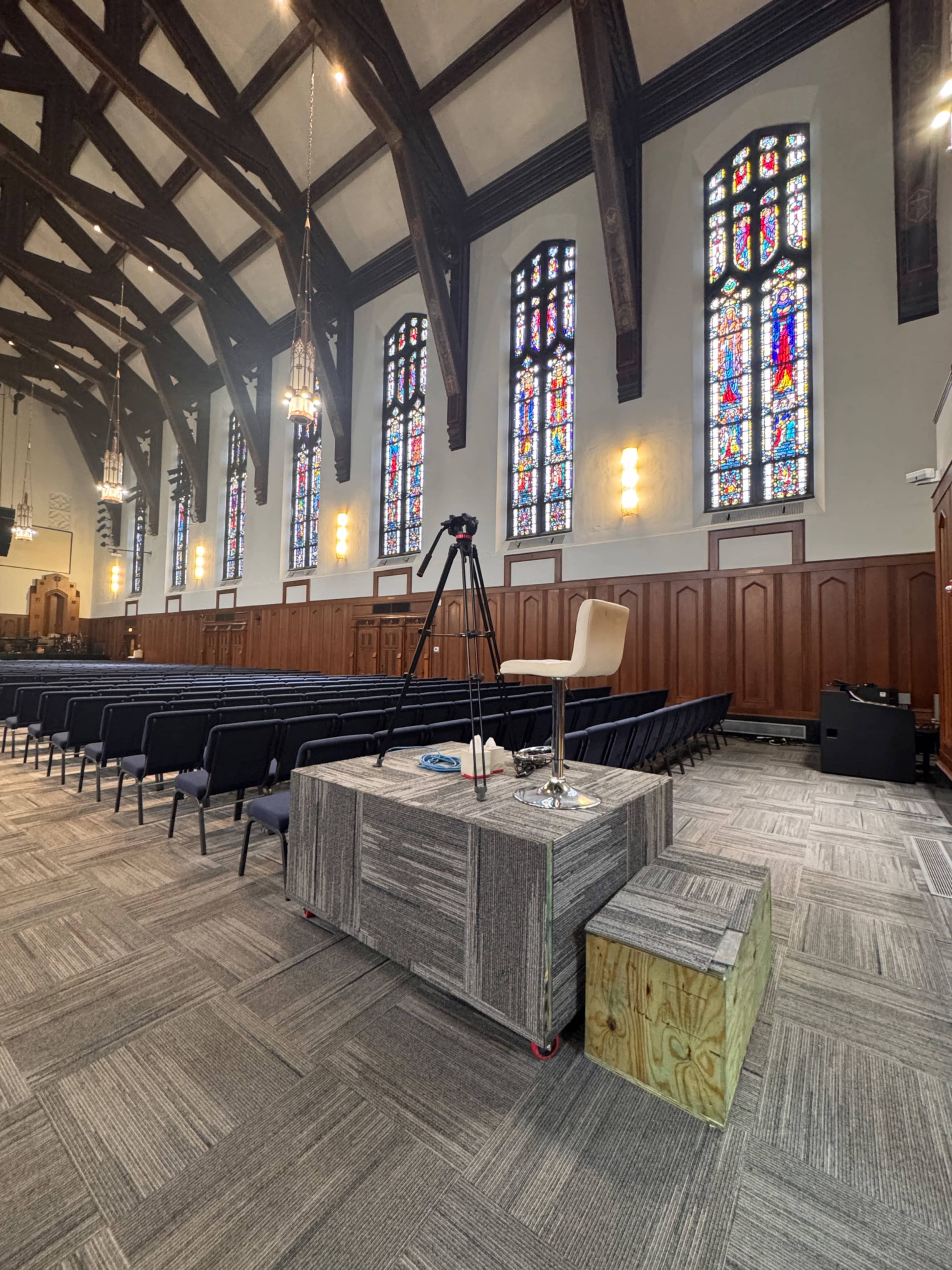 The image shows a church interior with high wooden beams, stained glass windows, rows of chairs, and a raised platform with a chair and camera equipment.