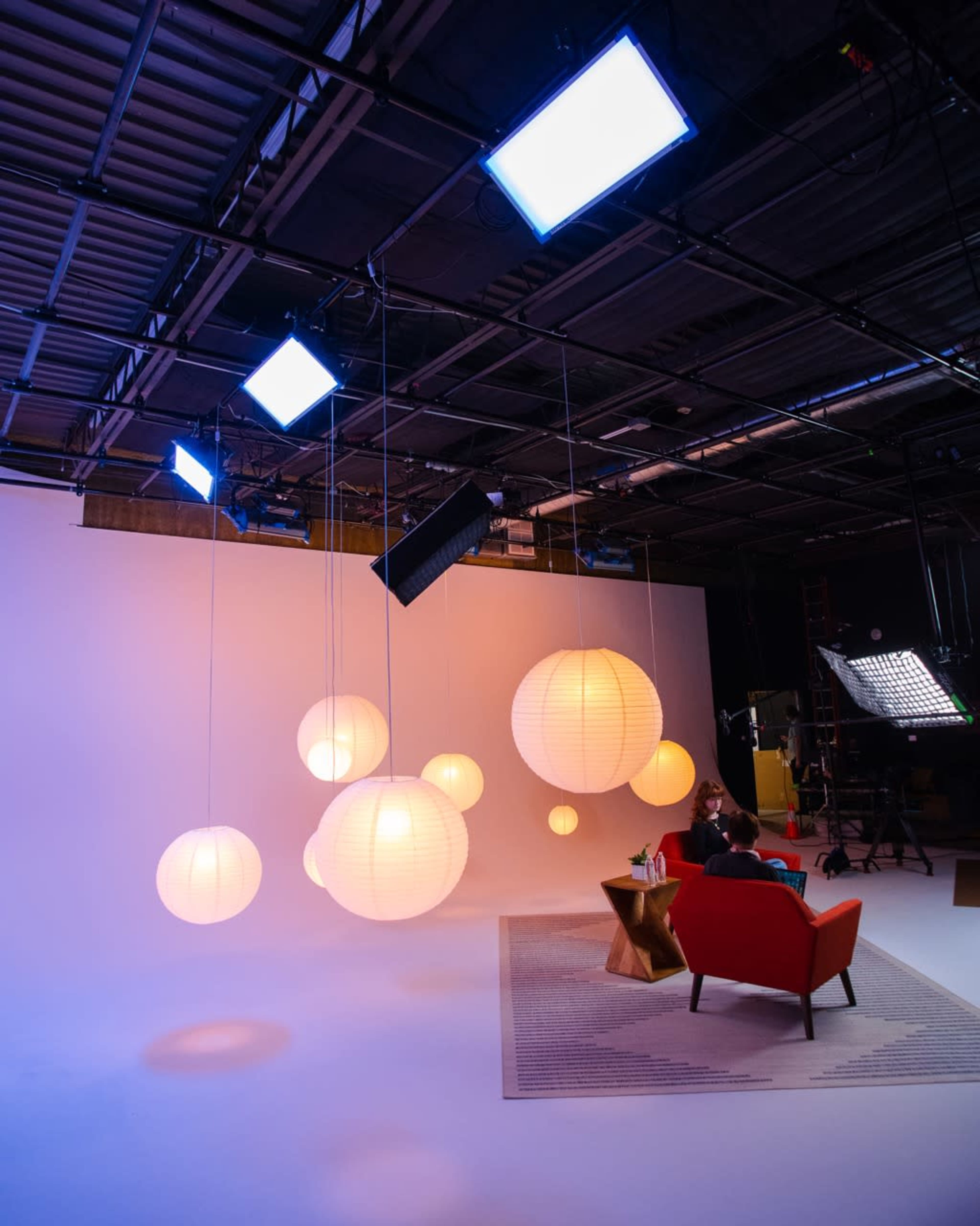 A studio setup features hanging paper lanterns, soft lighting, and a red armchair on a patterned rug against a gradient background.