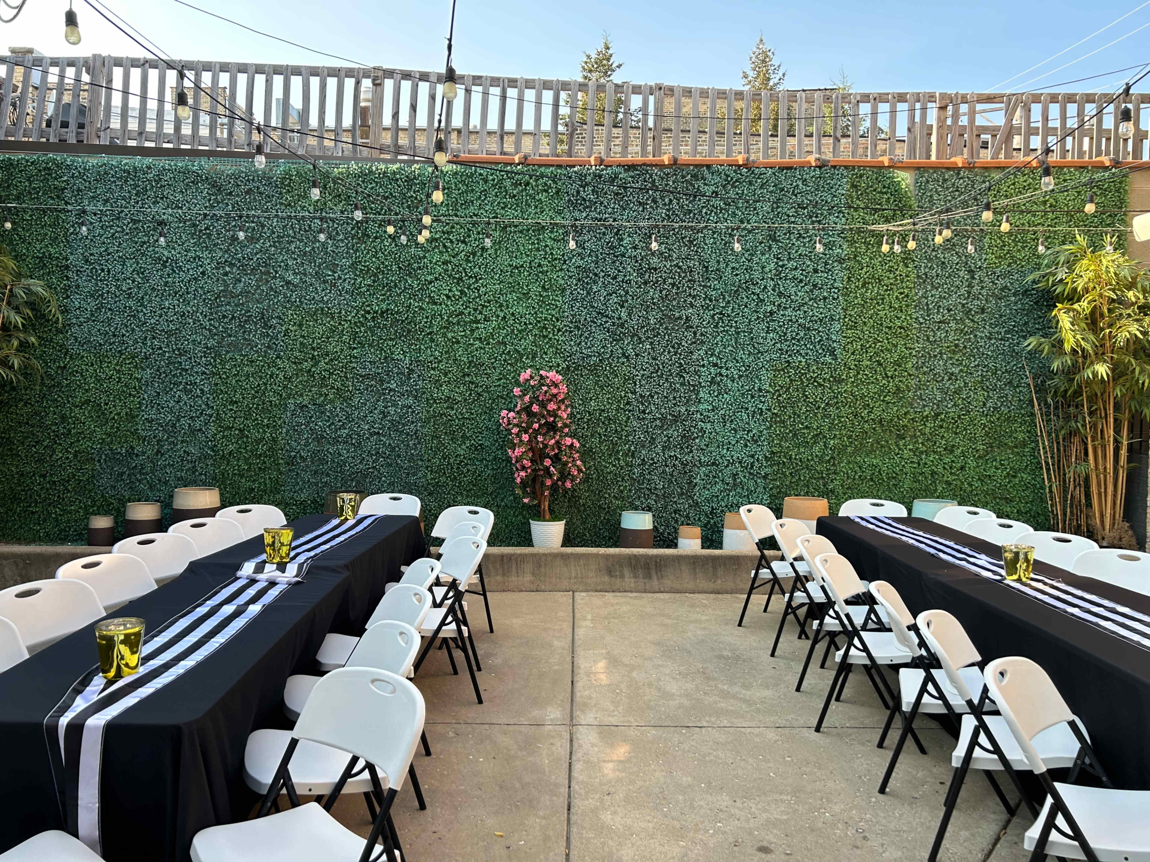 The image shows an outdoor space set up for an event, featuring several rows of folding chairs and tables covered with black cloth, alongside a vibrant green wall and hanging string lights.