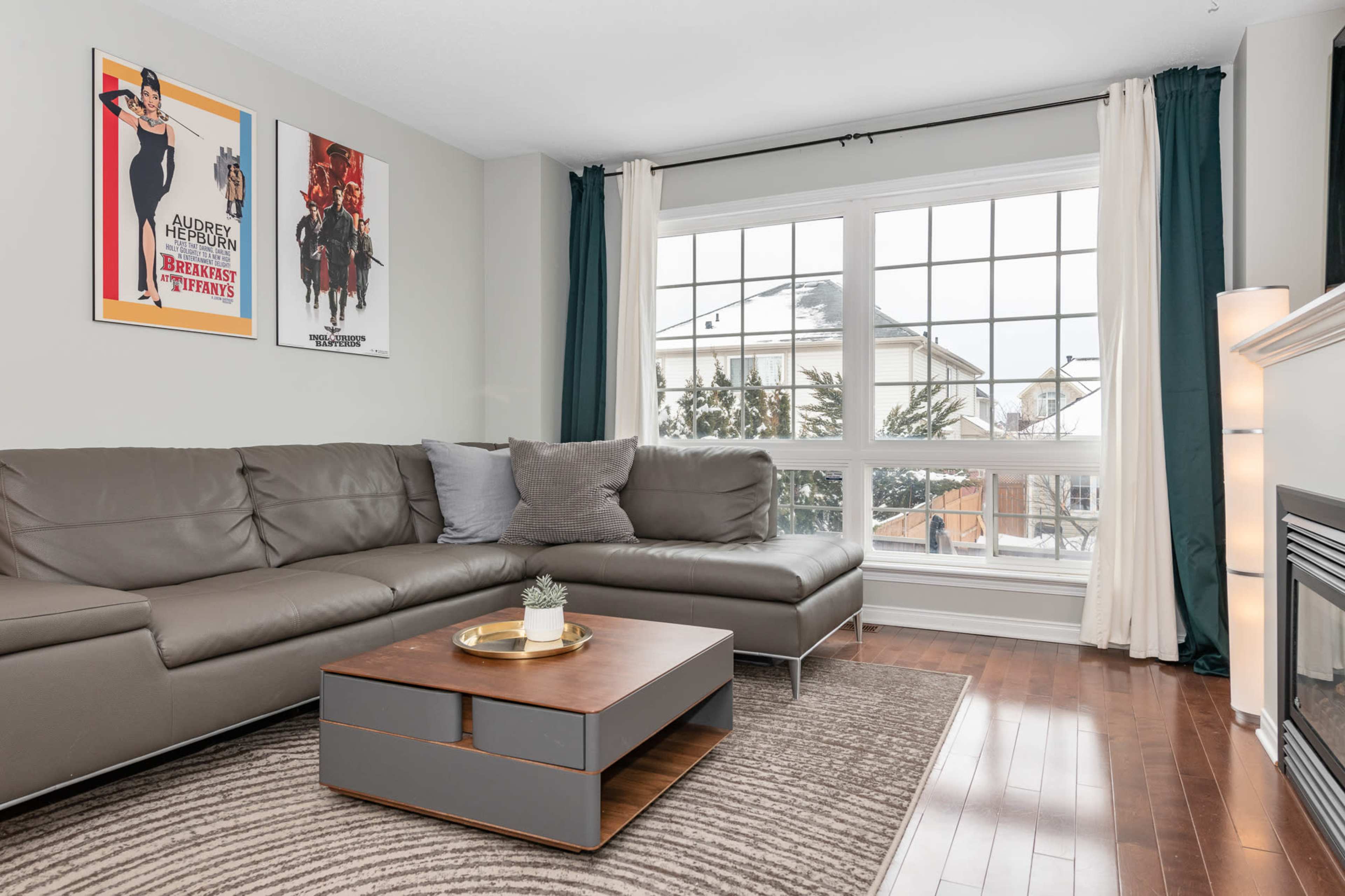 The living room features a gray sectional sofa, a wooden coffee table, and two framed movie posters on the wall, with large windows providing natural light.