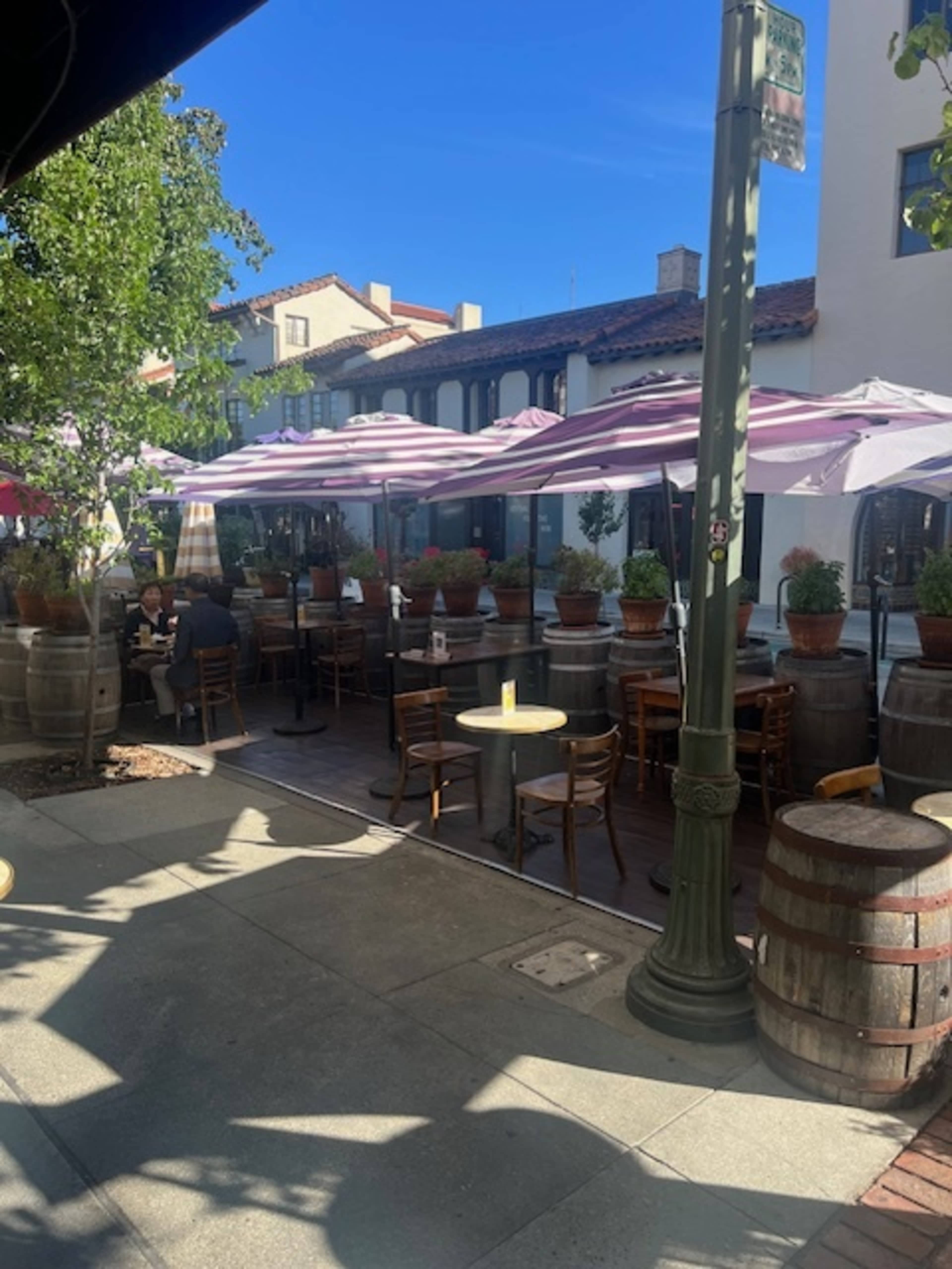 The image shows an outdoor restaurant patio with wooden tables, chairs, and purple umbrellas surrounded by potted plants and barrels, under a clear blue sky.